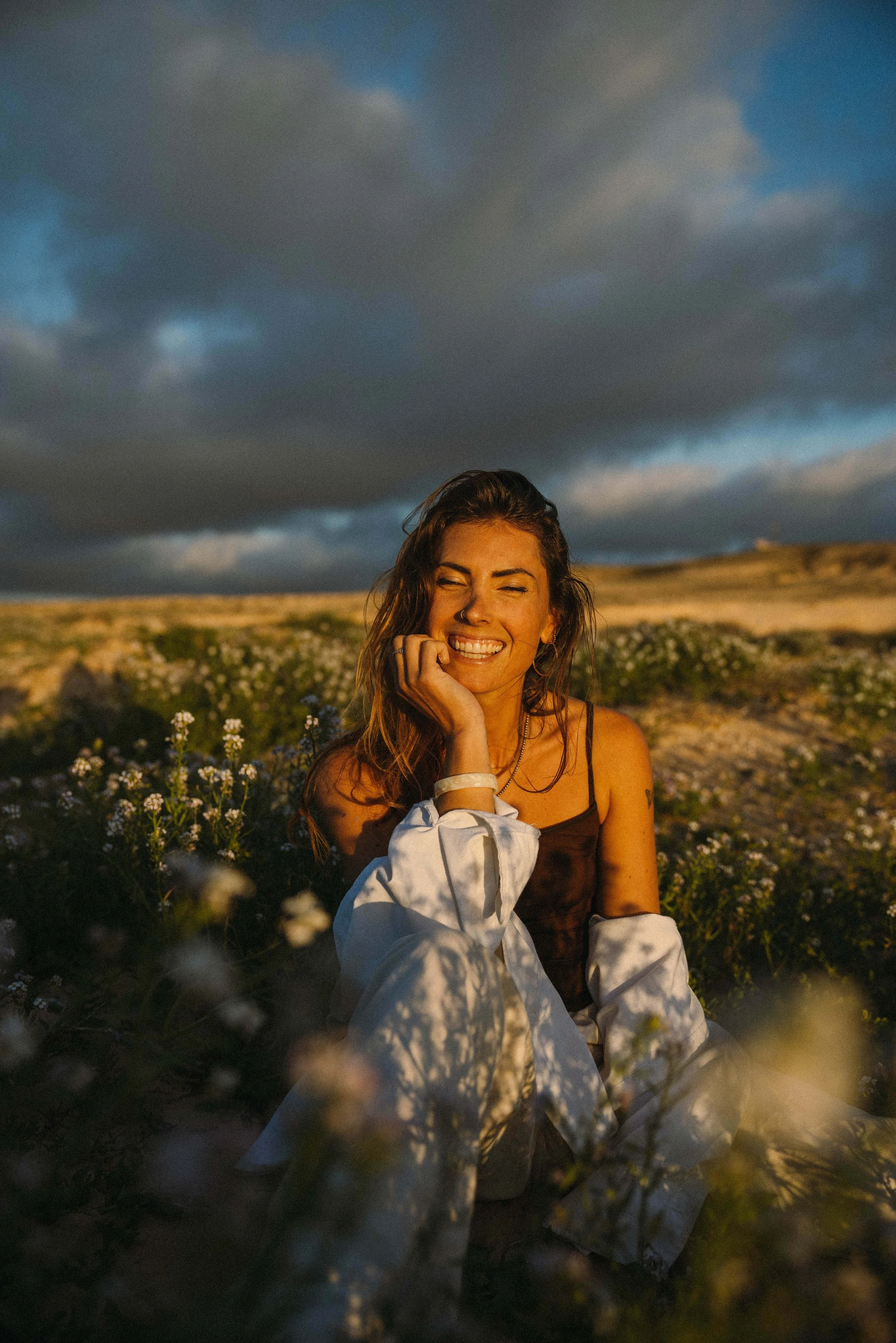 Eine lachende Frau sitzt in einem Feld mit Blumen bei Sonnenuntergang, dunkle Wolken am Himmel im Hintergrund.