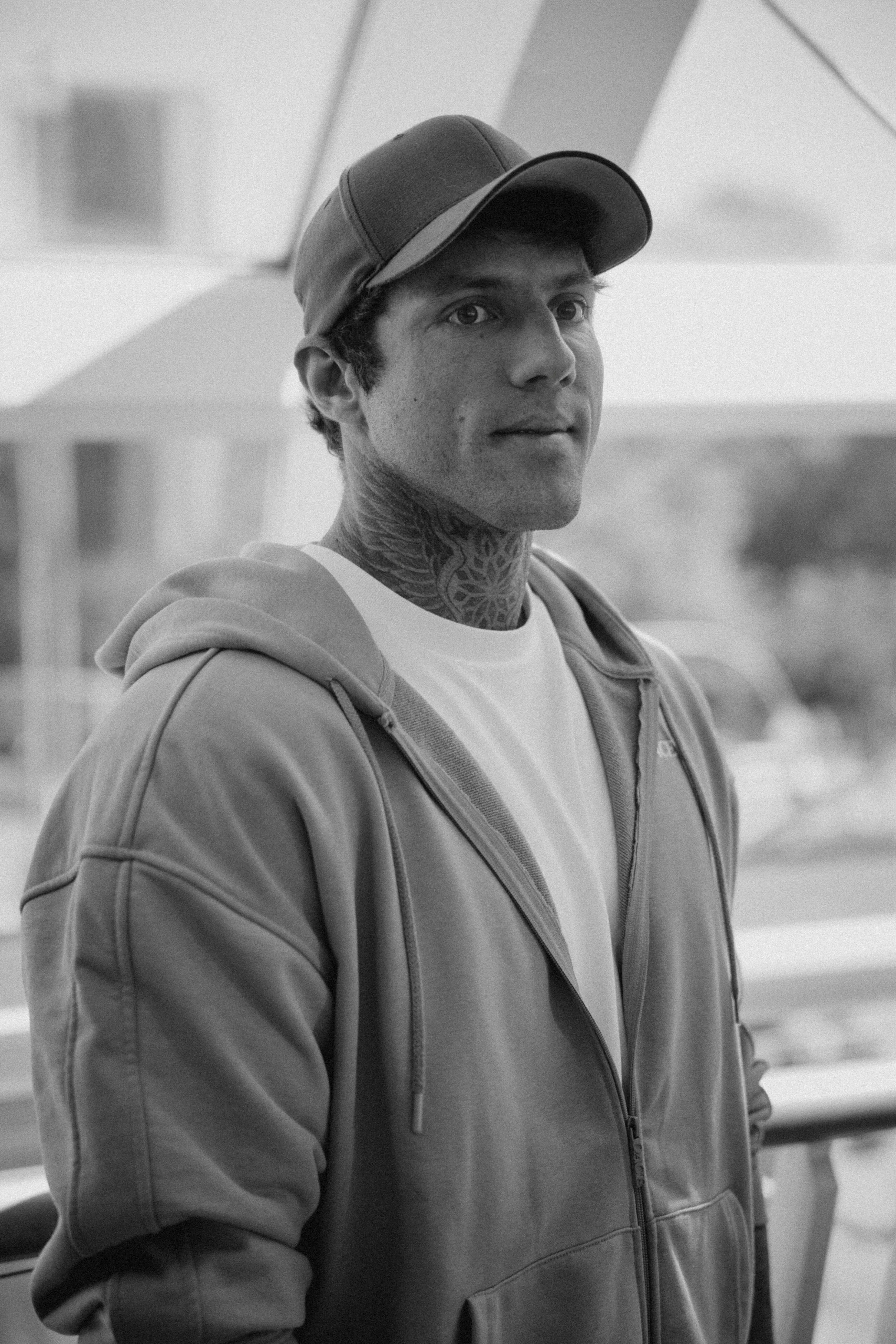 Schwarz-weiß-Foto eines jungen Mannes mit tätowiertem Hals, schwarzer Kappe und Hoodie, der in die Kamera schaut.