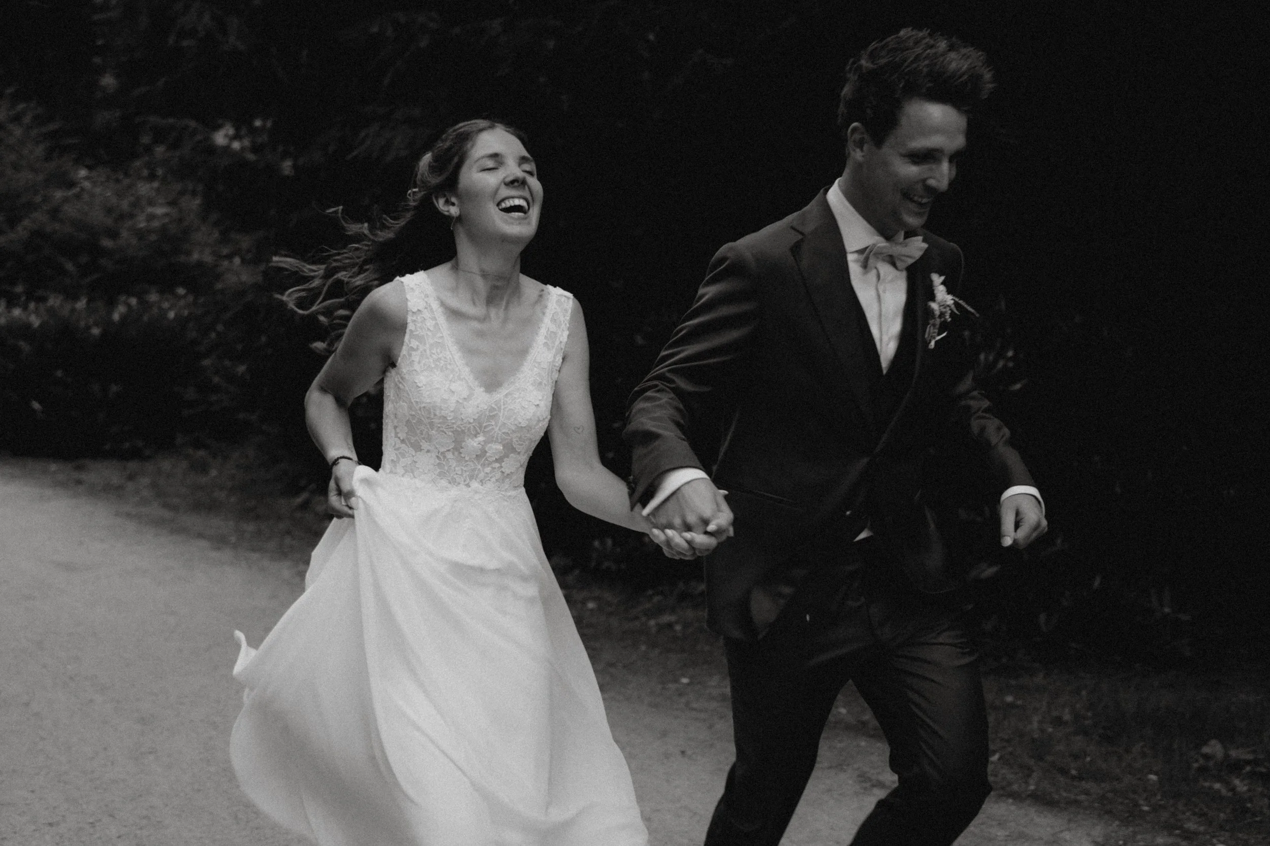 Ein glückliches Brautpaar läuft Hand in Hand auf einer dunklen Straße, beide lachen und tragen formelle Kleidung, die Frau trägt ein weißes Hochzeitskleid, der Mann einen schwarzen Anzug mit Fliege.