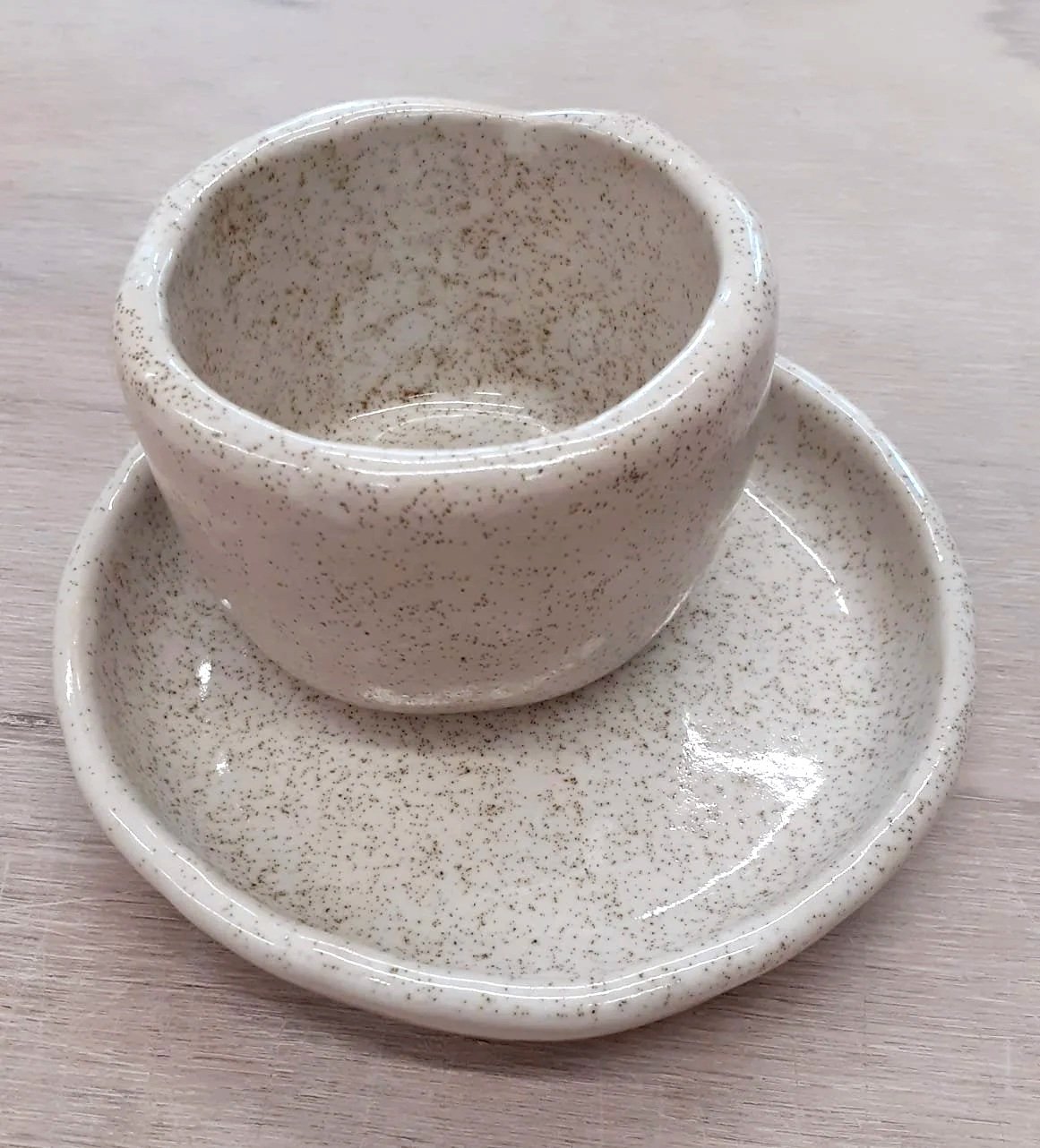 Poteries réalisées à la main et représentant un petit bol posé sur une soucoupe, les deux objets étant émaillés en blanc moucheté de noir.