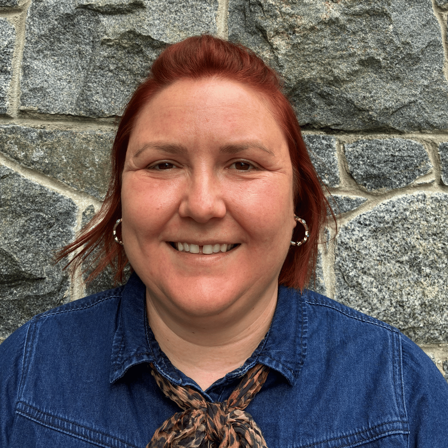 Close-up of a woman with shoulder-length blonde hair, smiling, in an office setting with desks and computers in the background.