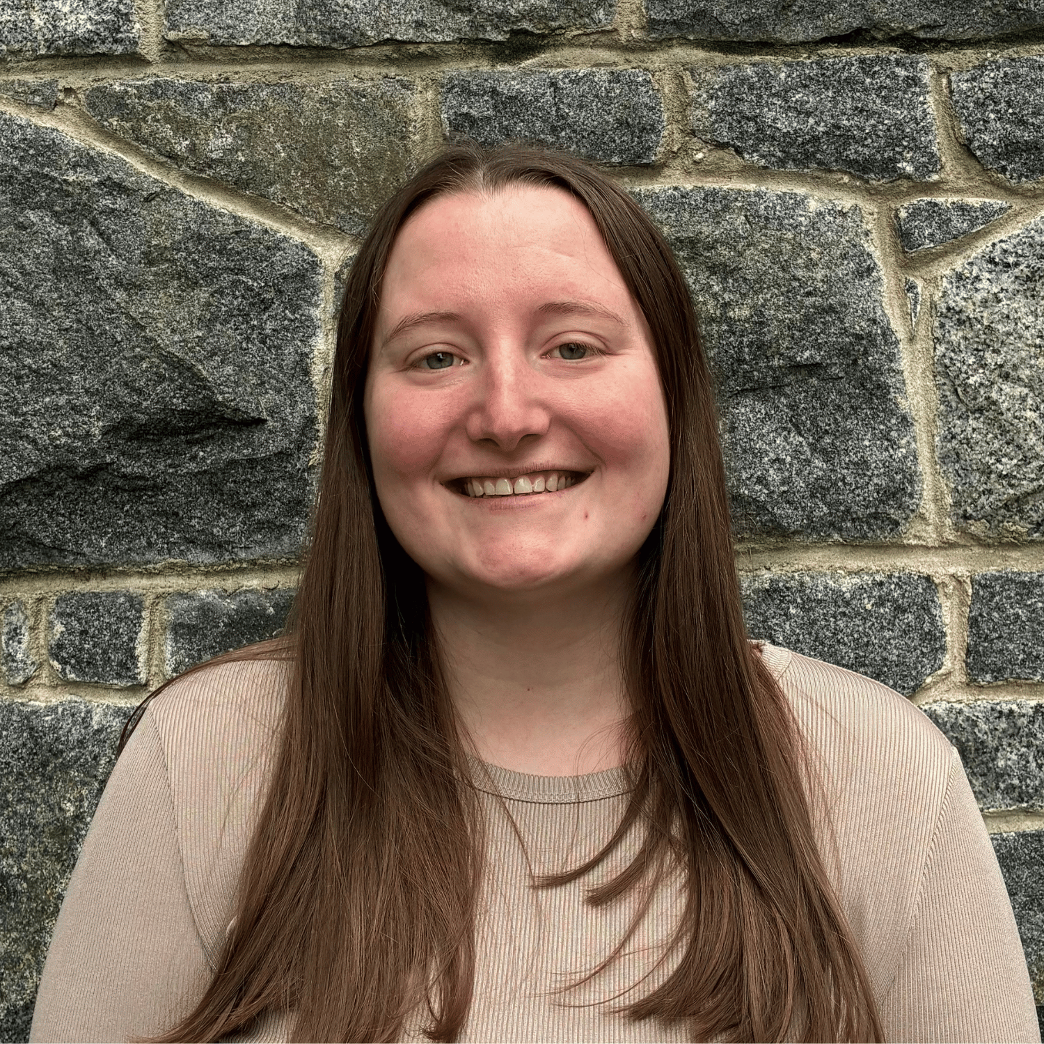 Headshot of a young woman with long red hair smiling, set against a blurred background of an office classroom with desks and chairs.
