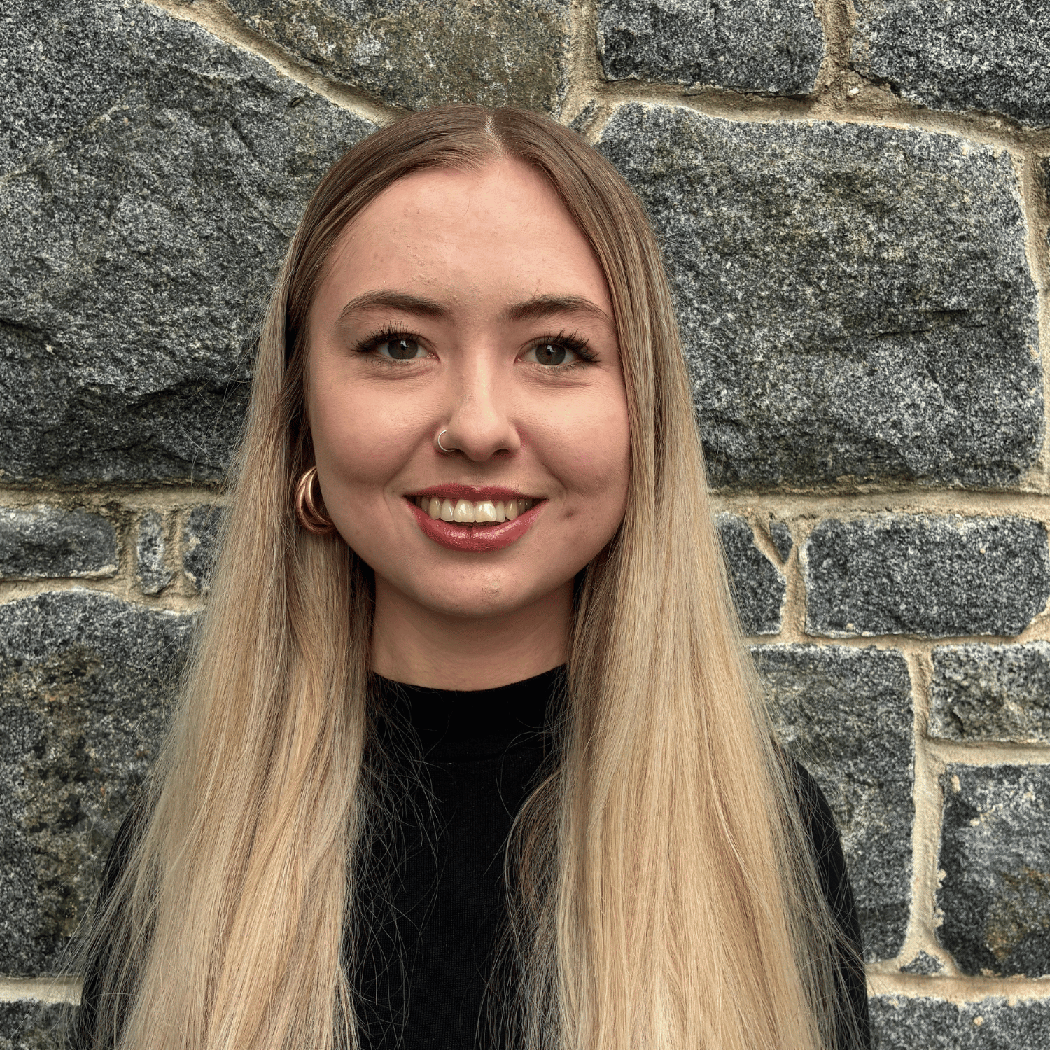 Young woman with blonde hair, wearing white earbuds and a white top under a dark cardigan, smiling in an office setting.
