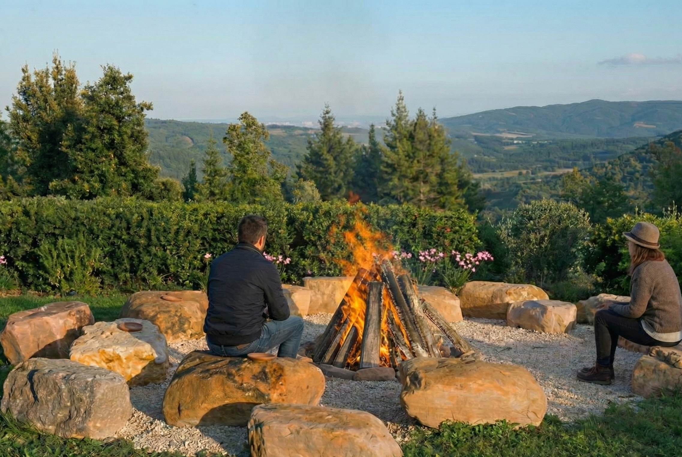 Two people sitting around a campfire outdoors in a scenic, hilly landscape with trees and mountains in the background, surrounded by large rocks and greenery.