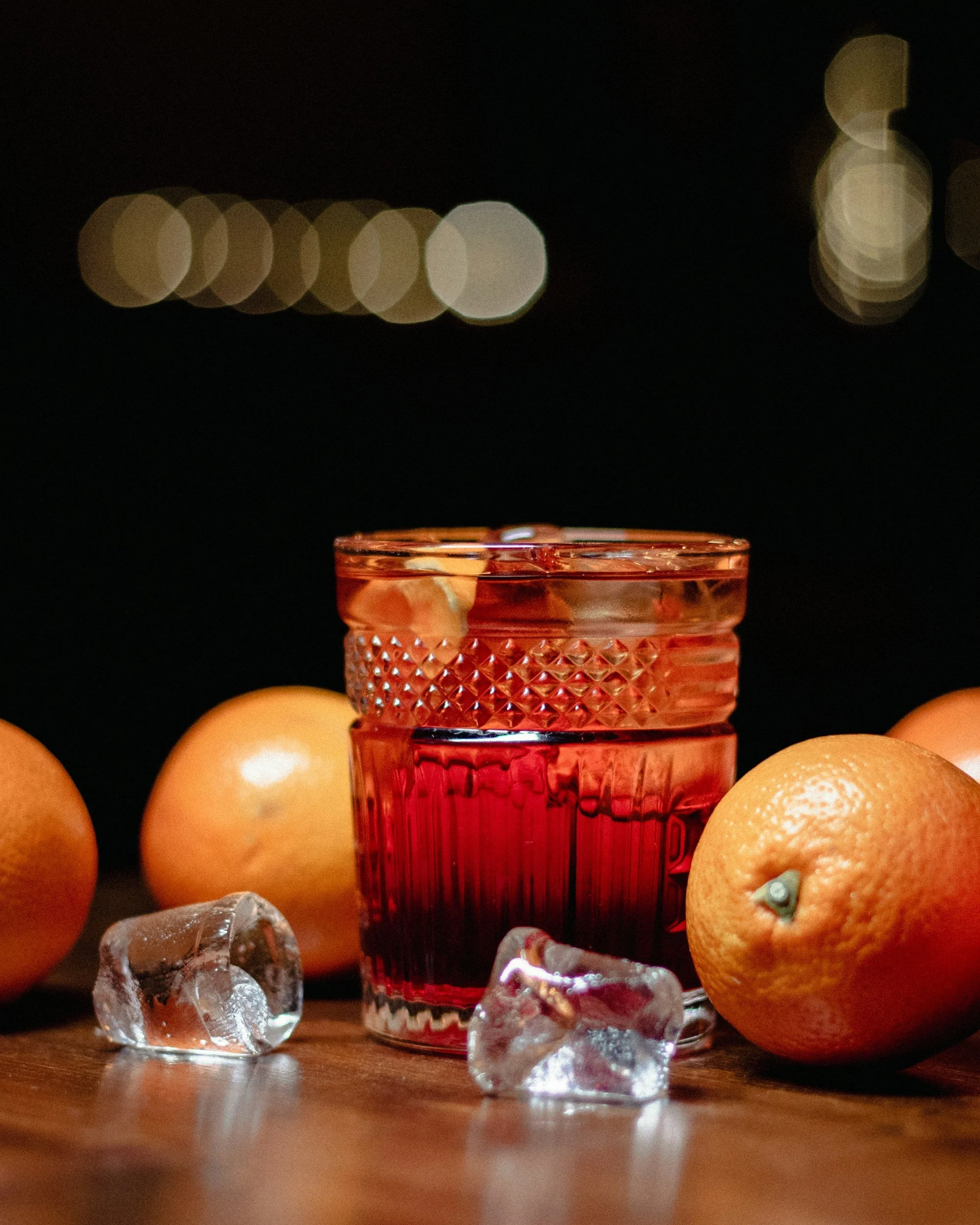 A pink cocktail glass with a textured pattern filled with a red drink, surrounded by oranges and ice cubes on a wooden surface, with blurred city lights in the background.