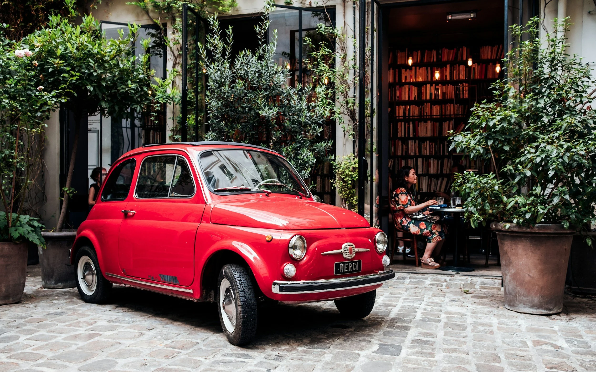 Red vintage Fiat parked outside a café with a person working on a laptop inside, surrounded by greenery and large potted plants.