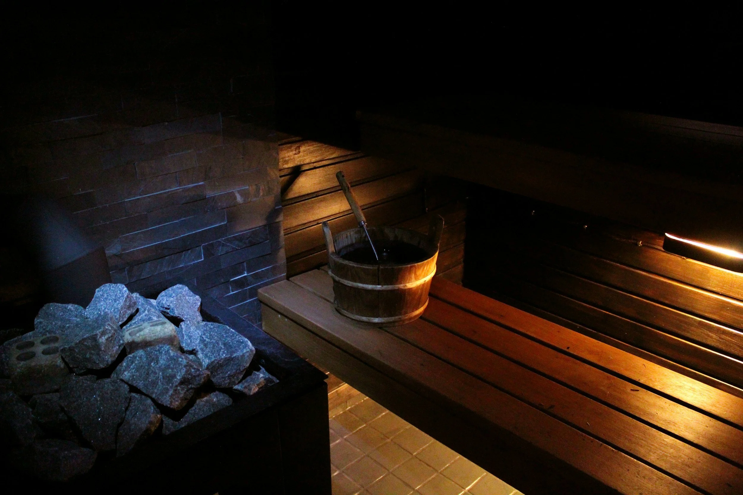 Dimly lit sauna room with wooden benches, a bucket of water with a ladle, and a heater with stones.