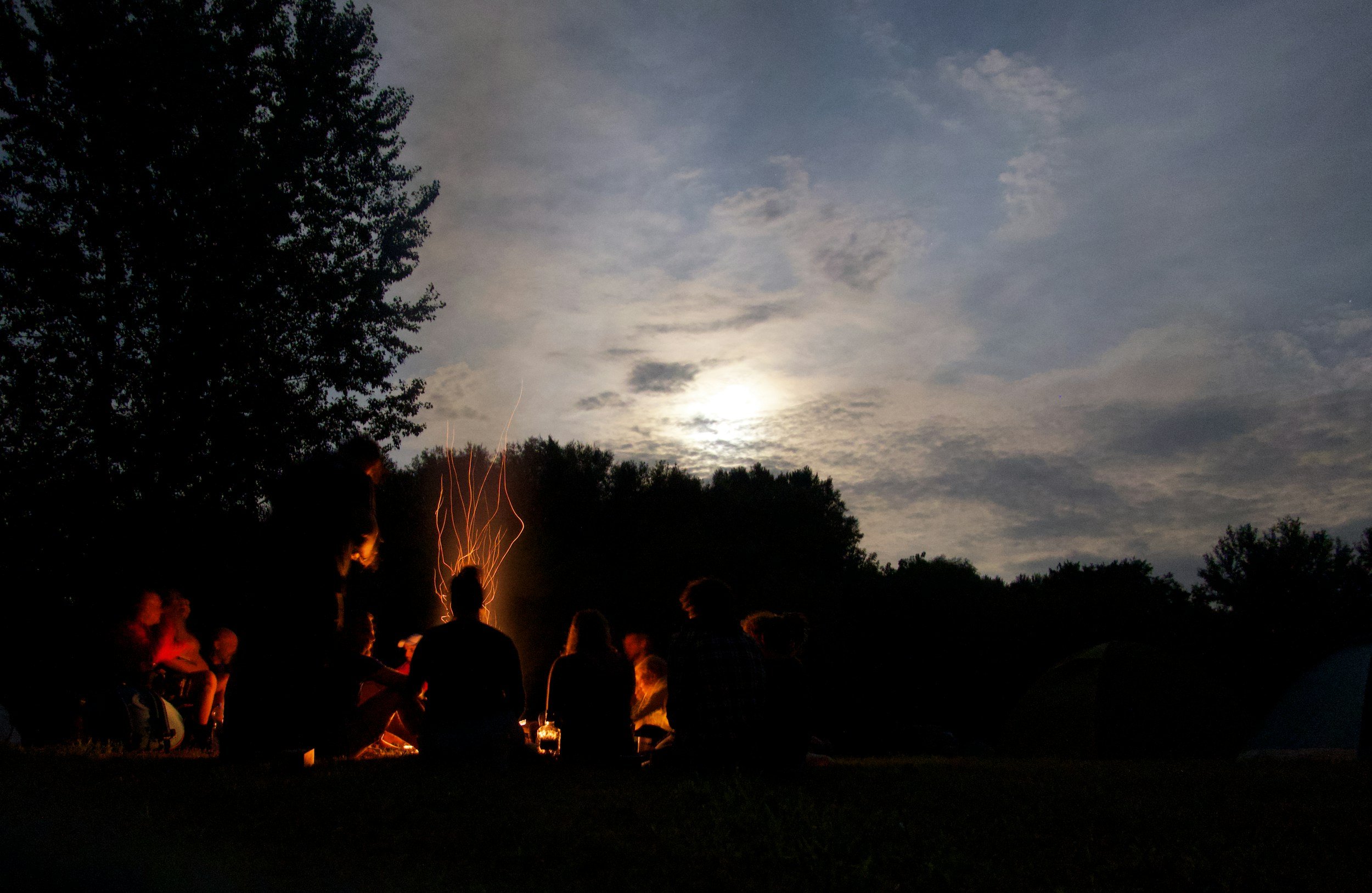 People sitting around a campfire at night under a partly cloudy sky with a bright moon.