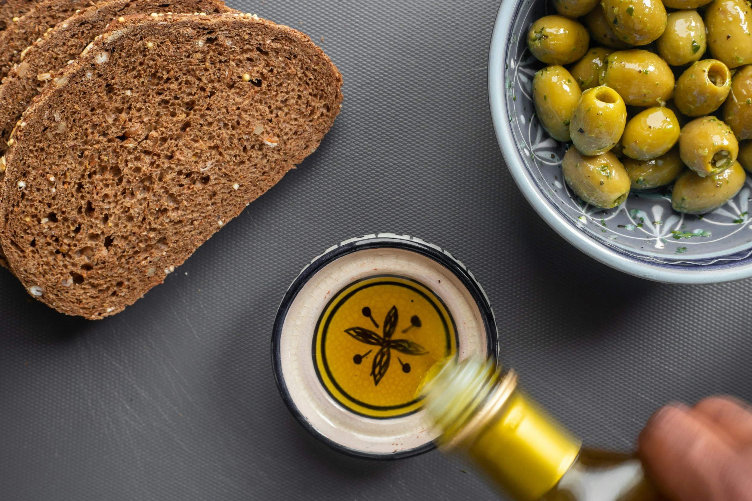 A loaf of sliced brown bread, a bowl of green olives, and a small dish of olive oil on a gray surface.
