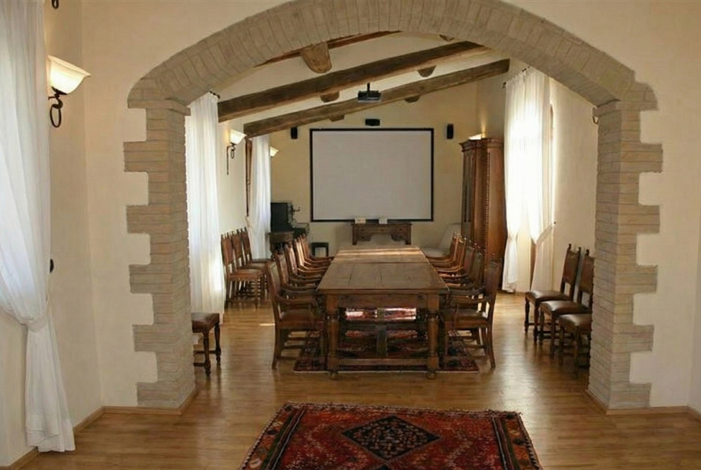 A spacious conference room with a long wooden table surrounded by 12 chairs, a large projection screen on the wall, and windows with white curtains letting in natural light. There are stone archways and wooden beams overhead, and a decorative rug on the wooden floor.