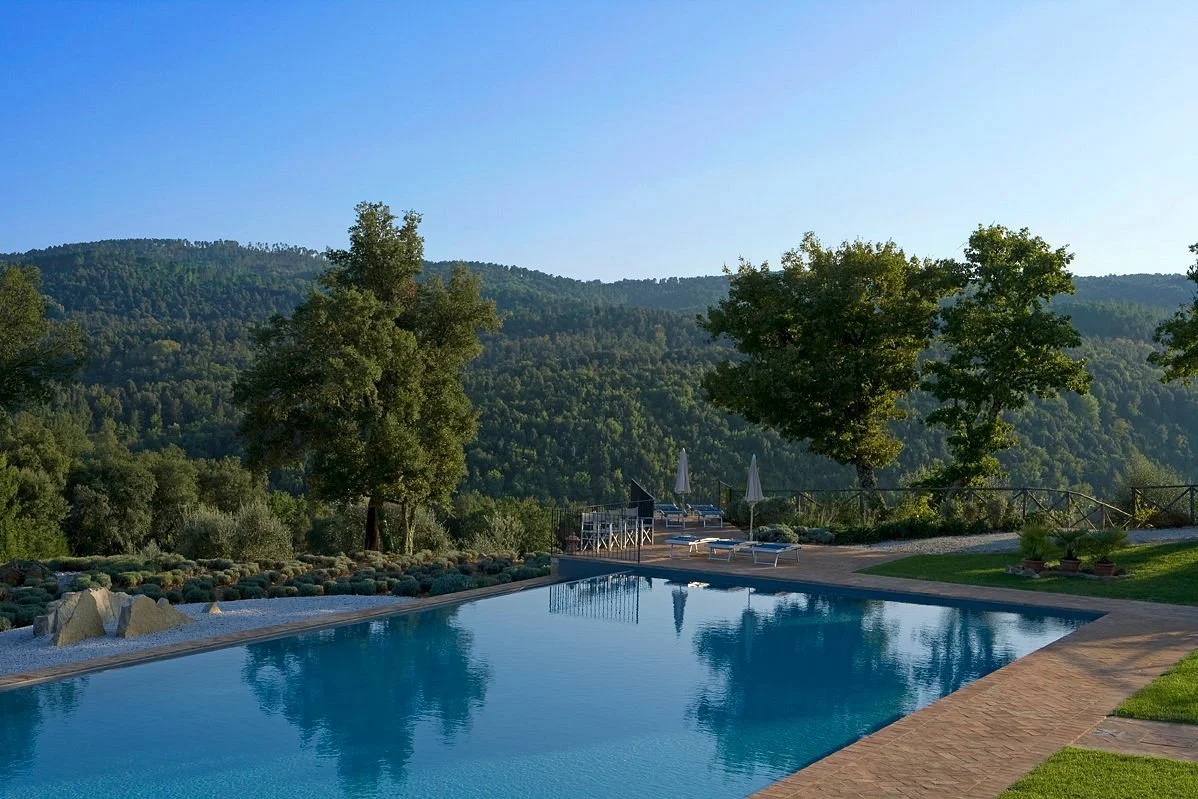 A peaceful outdoor swimming pool area with mountain views, surrounded by trees, lounge chairs, and umbrellas in a natural setting.