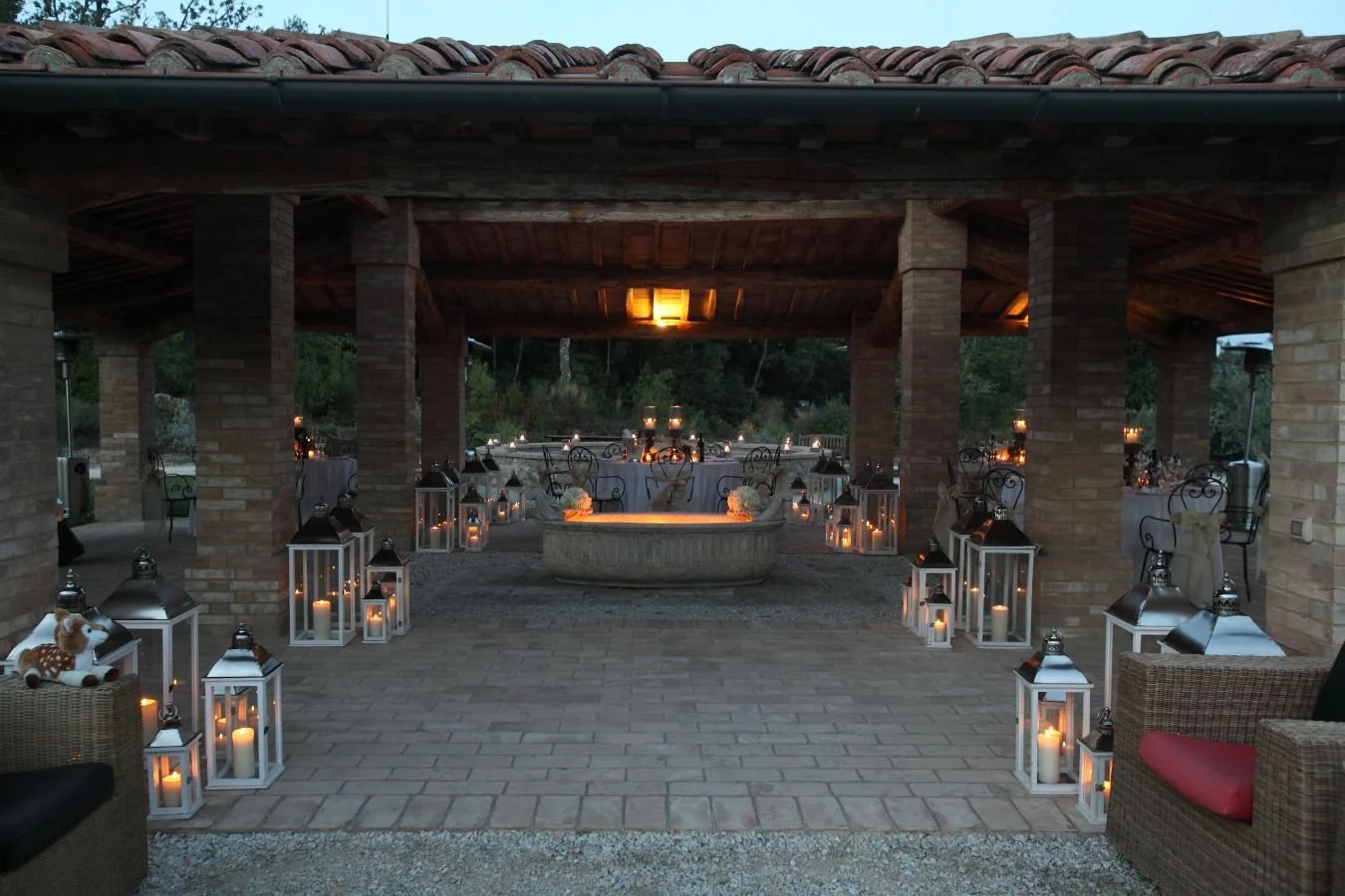 Decorated outdoor patio with lanterns, candles, and elegant tables set for an evening event under a covered brick structure.