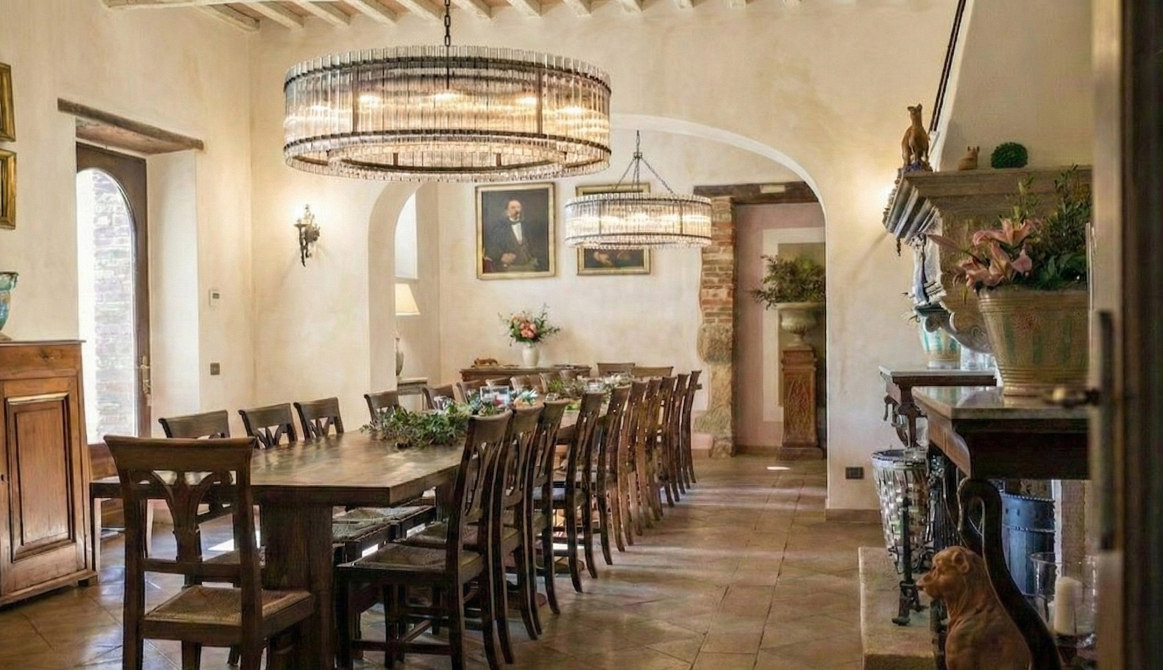 Elegant dining room with a long wooden table surrounded by chairs, chandelier lighting, and decorative elements like paintings, vases, and figurines. A dog is sitting near the fireplace.