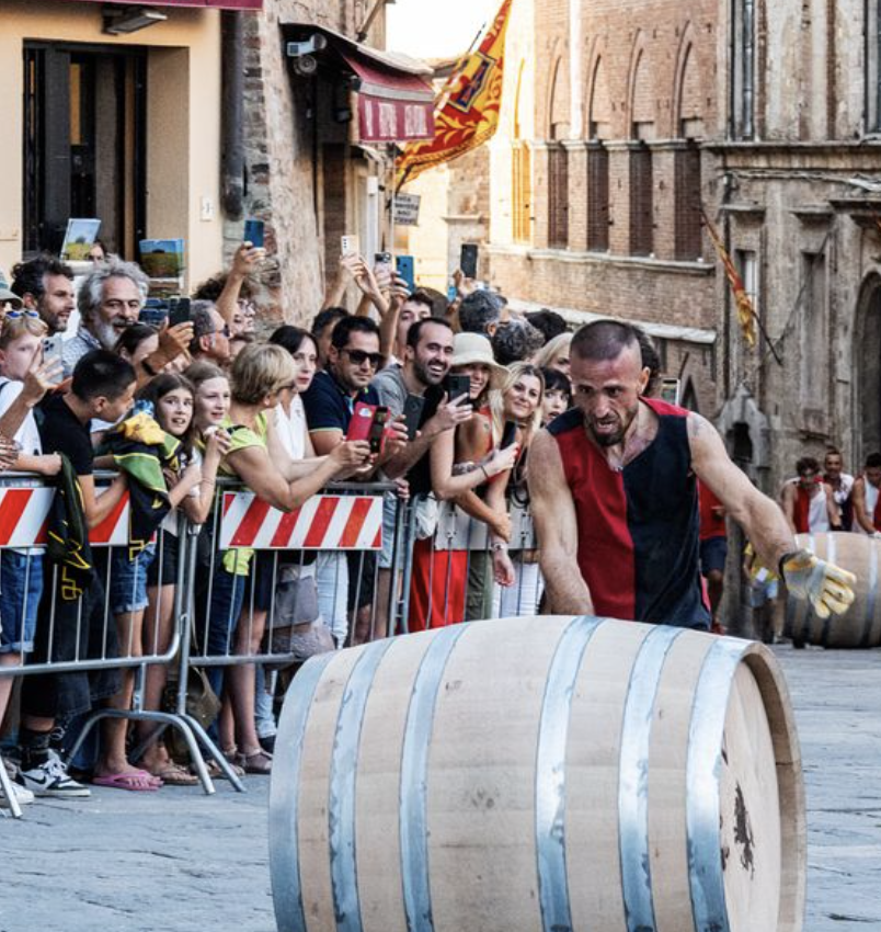 A man participating in a barrel rolling race during a street festival, with a crowd of onlookers behind a barricade watching and taking photos.