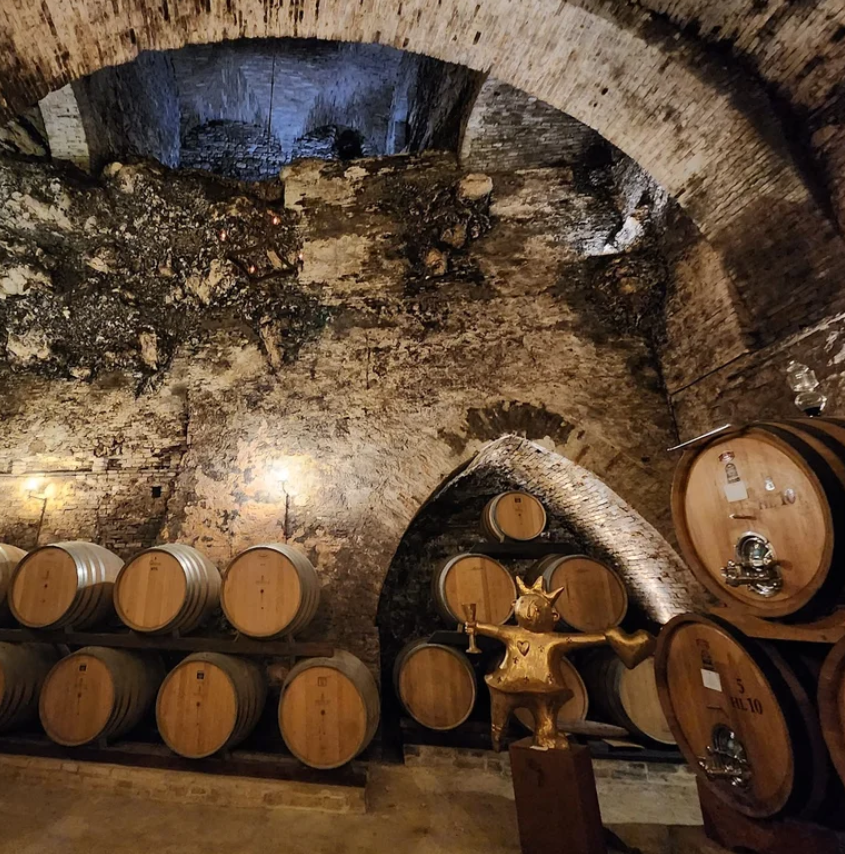 Wine barrels stacked on racks beneath a vaulted, brick ceiling in a cellar with exposed brick walls and dim lighting.