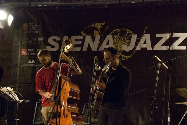 Two musicians playing jazz instruments on stage at a jazz festival with a black backdrop and the words 'SENA JAZZ' in large white letters.