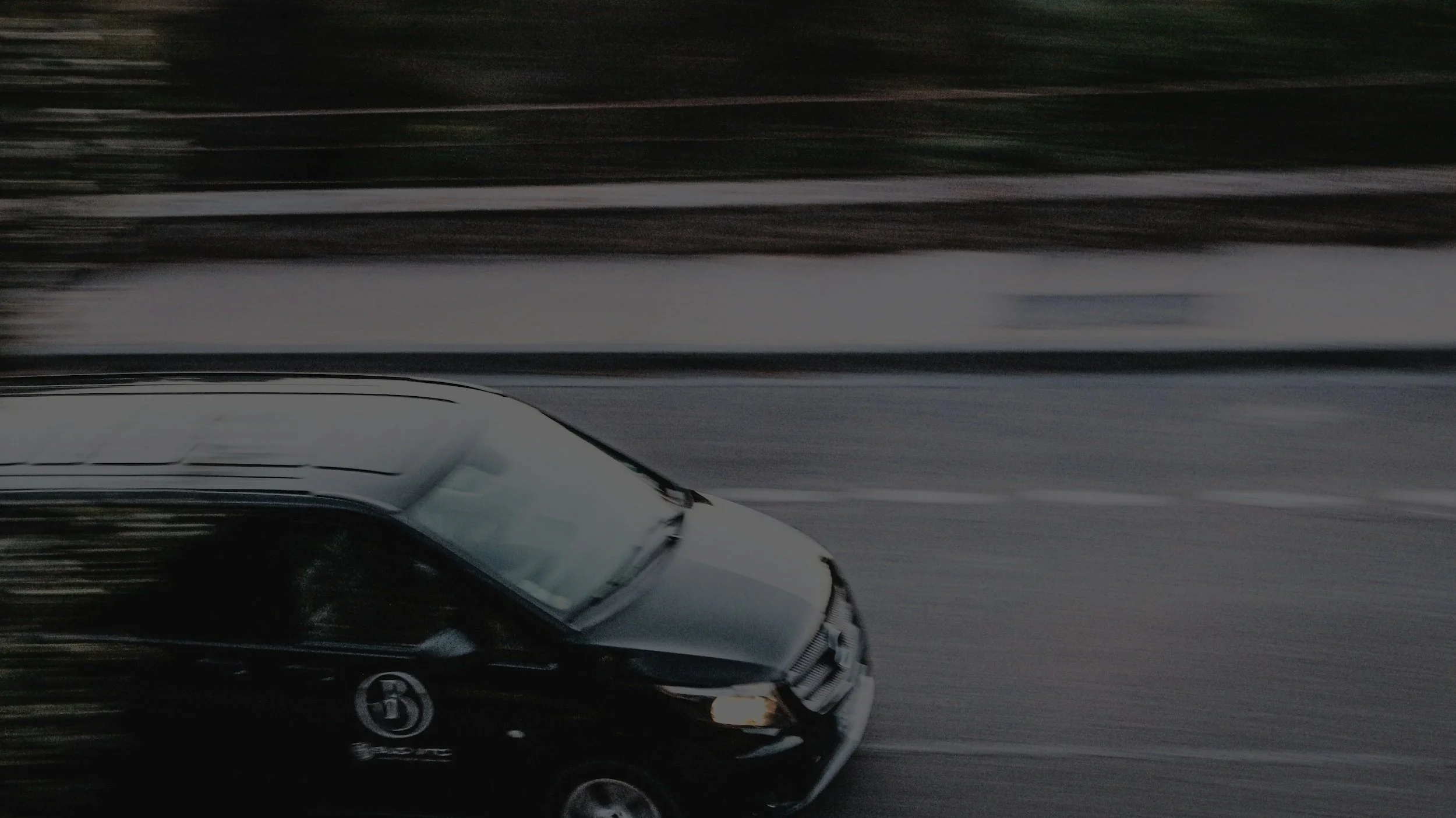 Moving black car with a logo on the front door driving on a highway surrounded by trees.