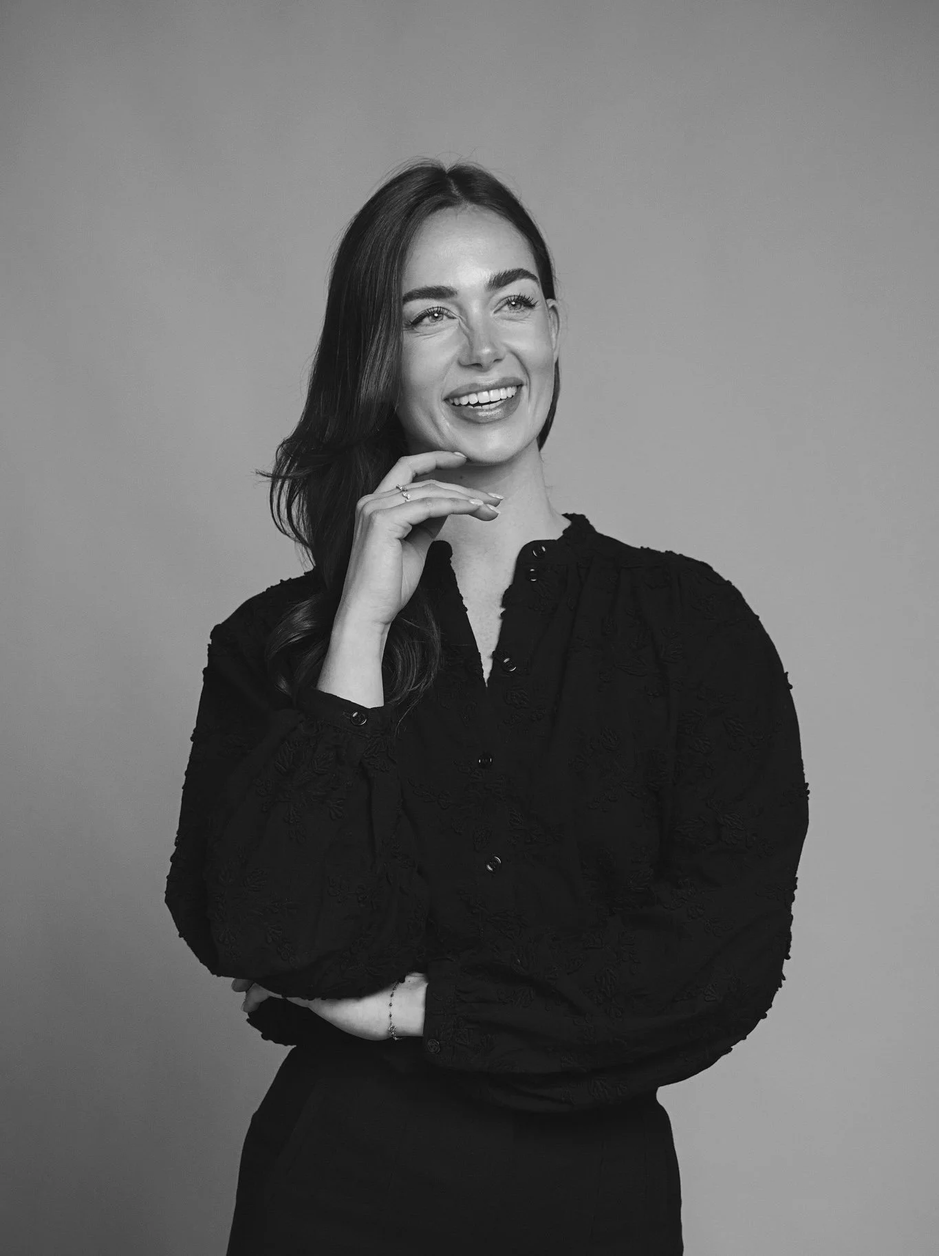 Black and white studio portrait of a smiling woman with wavy hair, wearing a dark embroidered blouse, standing against a plain background.