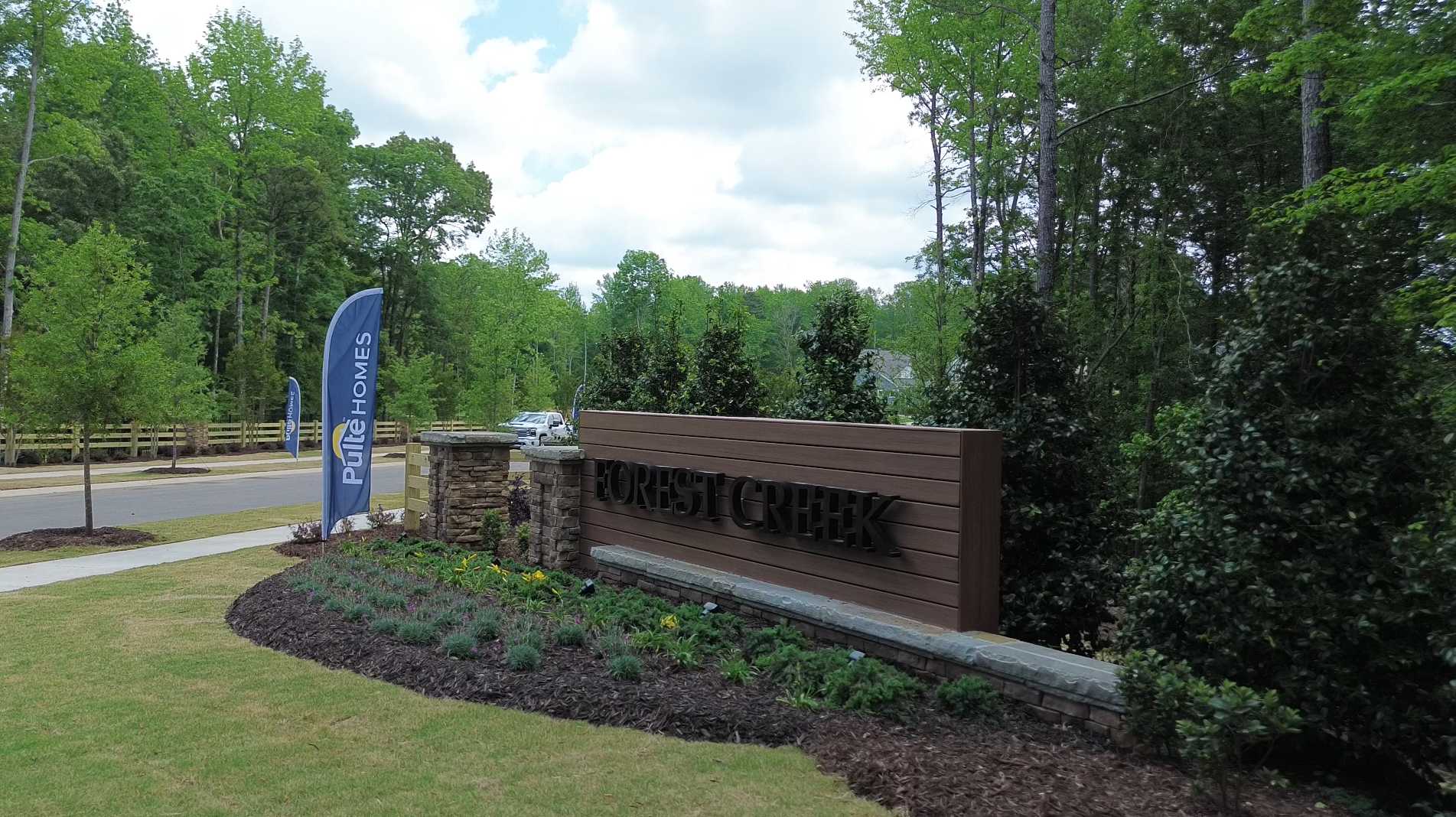 Entrance sign for the Forest Creek community, set in landscaped greenery with shrubs, flowers, and a stone pillar, alongside a road with Pure Homes banners.