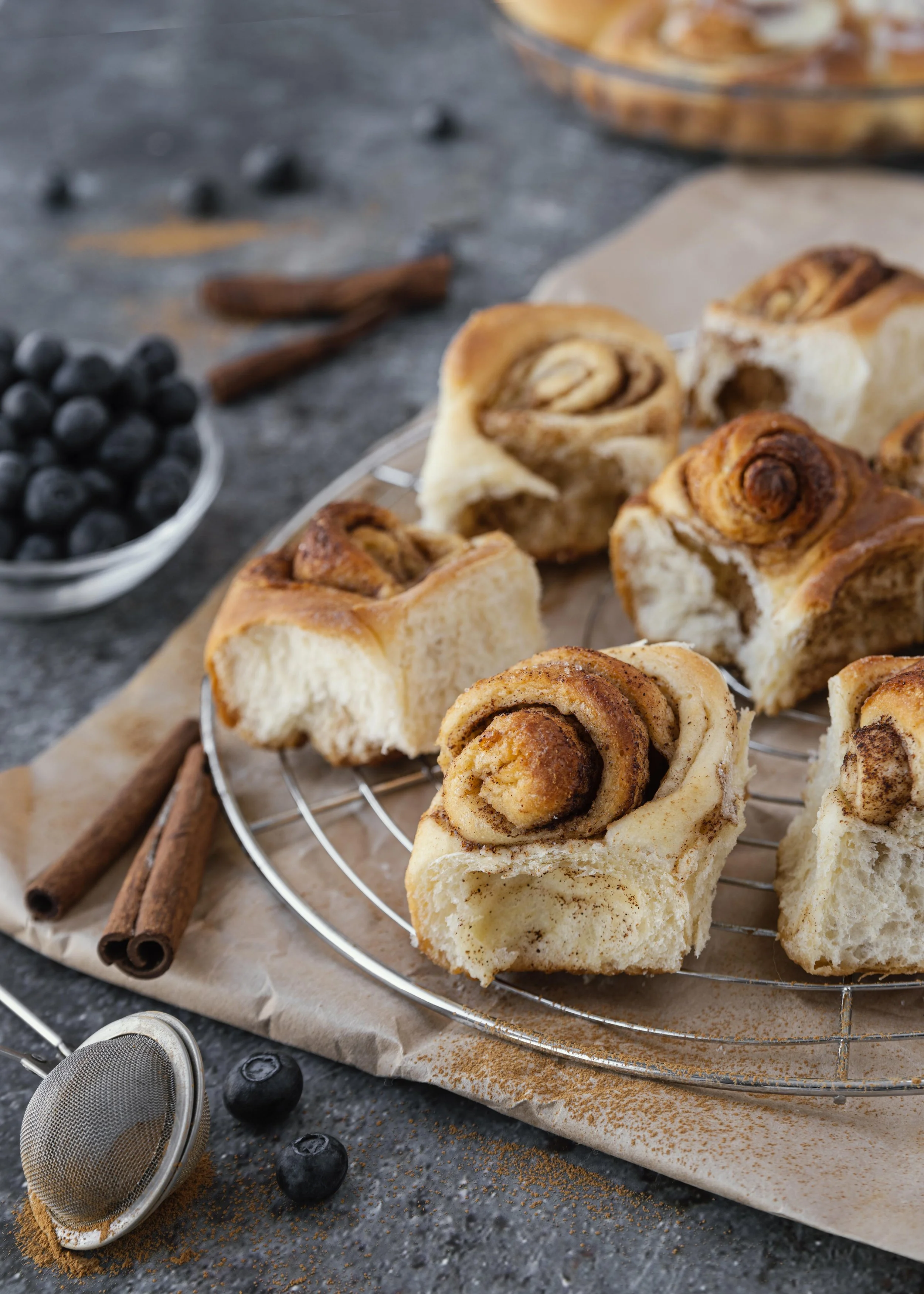 Scones med kanel og sukker, dekorert med kanelpulver, på et avkjølingsstativ. Det er friske blåbær, kanelstenger og et sølvstålssil i nærheten.