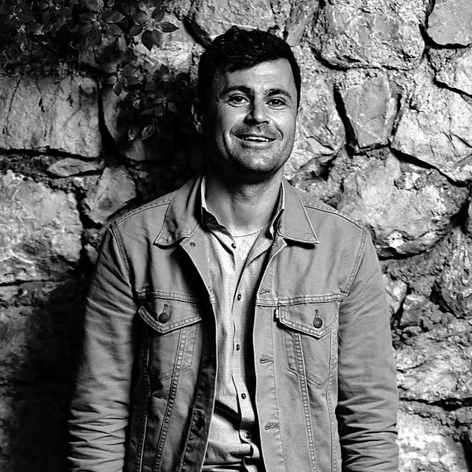 A smiling man wearing a denim jacket and a collared shirt standing against a stone wall.