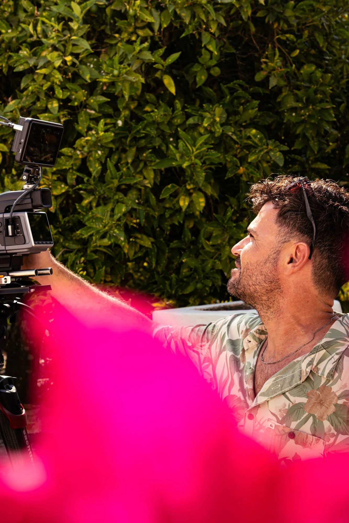 A man with curly hair and a beard wearing a Hawaiian shirt setting up filming equipment outdoors near green bushes, with pink flowers in the foreground.