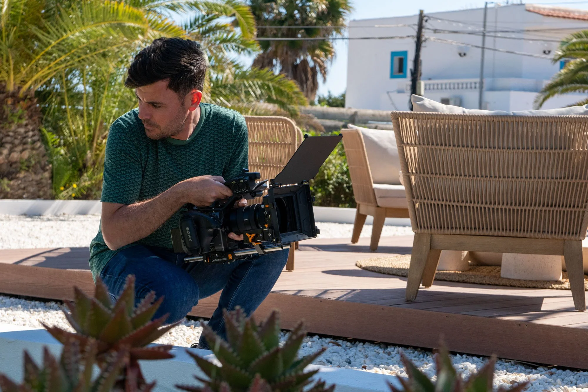 Man operating a professional video camera outdoors on a sunny day, with outdoor furniture and greenery in the background.