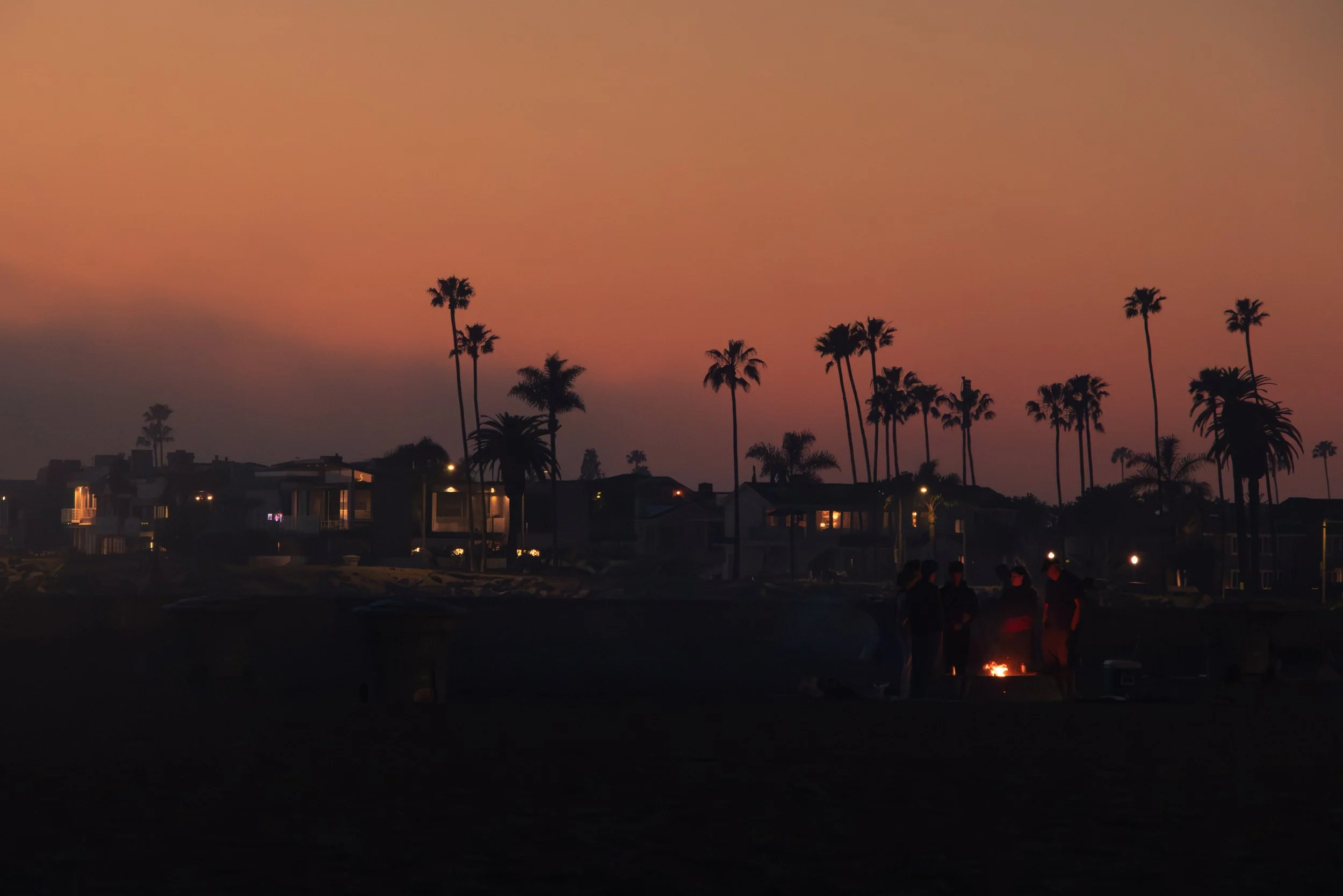 People gathered around a small fire during sunset, with palm trees and houses in the background.