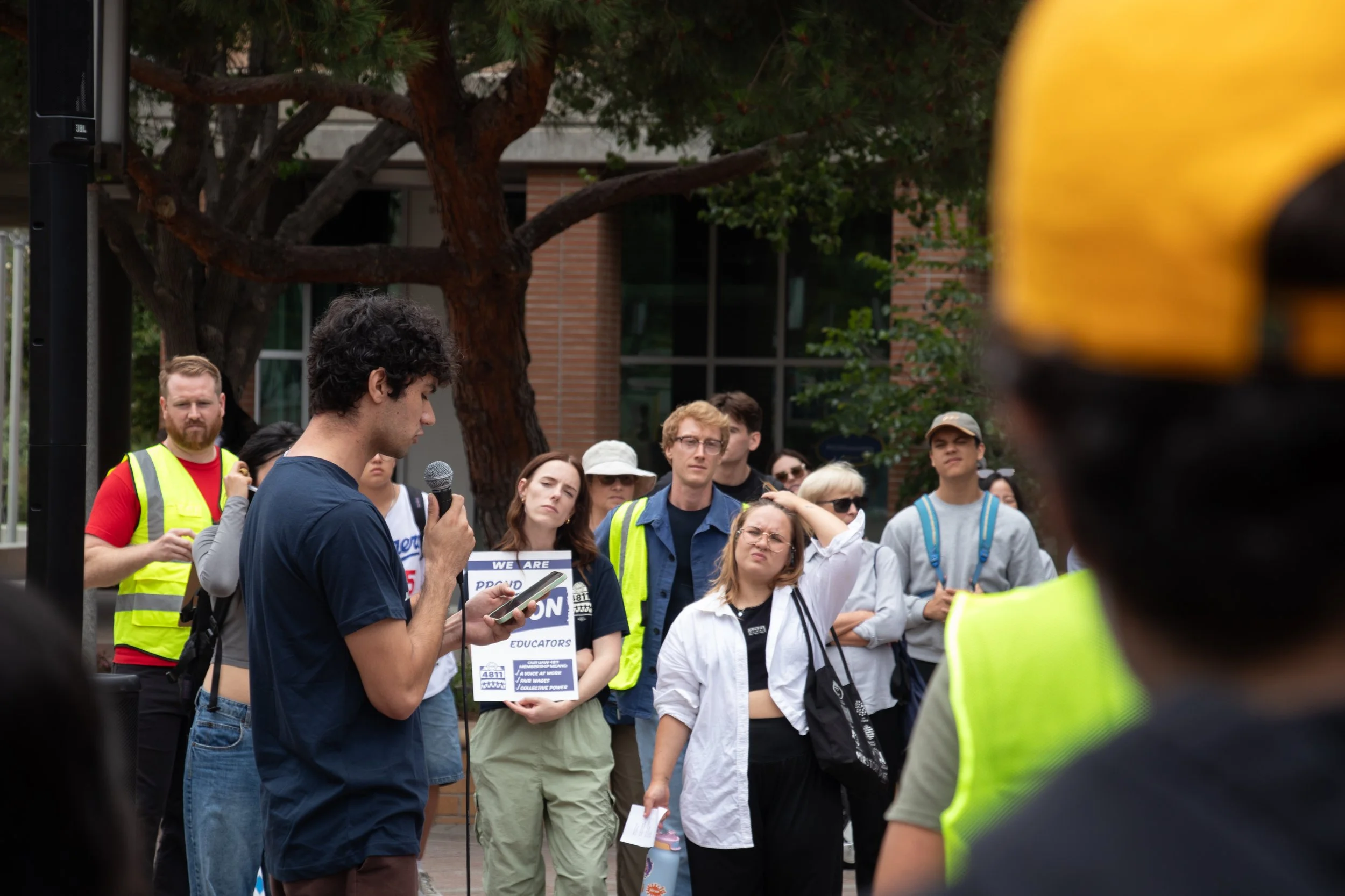 A young man stands reading from a phone while holding a microphone during a rally or protest. Several people around him are listening, some holding signs, with brick and glass building in the background and trees.