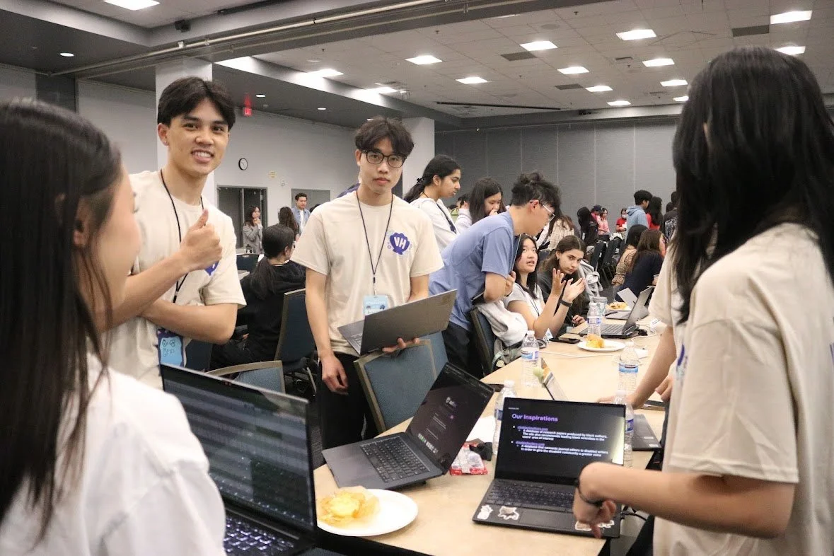 Group of young people gathered around a table with laptops, engaging in discussion during an event or workshop in a large conference room.