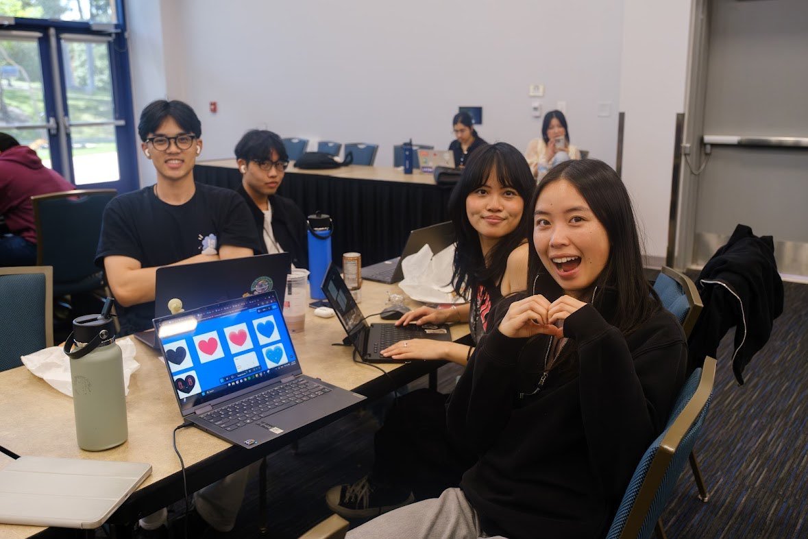 A group of five young people sitting around a table in a conference room, with laptops and drinks, smiling and making heart shapes with their hands.