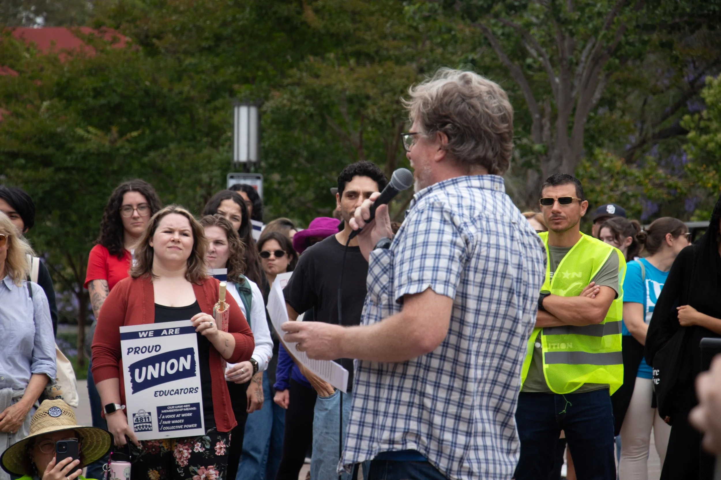 A man with glasses and a plaid shirt speaking into a microphone during a protest or rally, with a diverse group of people holding signs and listening attentively, in a park with trees in the background.