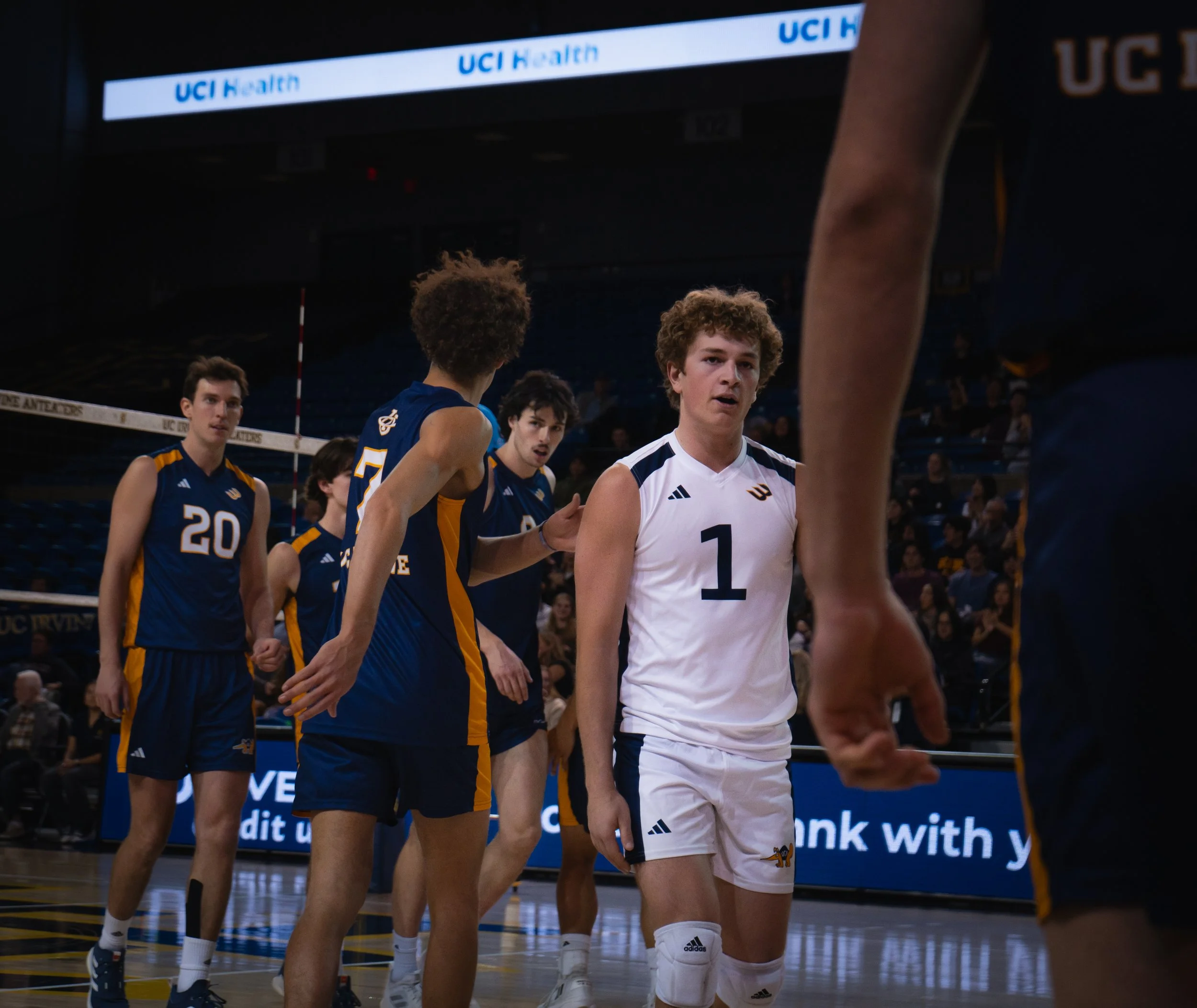 Young male volleyball players walking on the court, wearing blue and white uniforms, with an audience in the background at UCI Health arena.