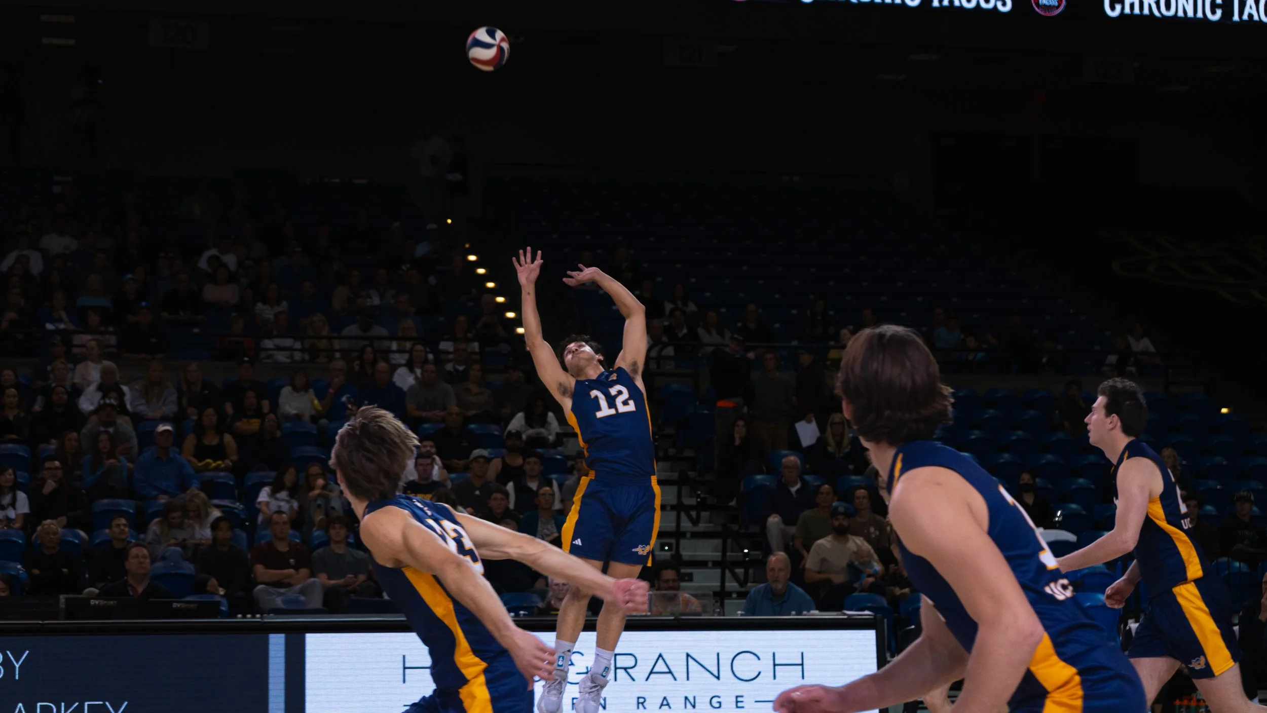 Indoor volleyball game with players in blue and yellow uniforms, one player jumping to spike the ball while others move to block or assist. Audience seated in the background.