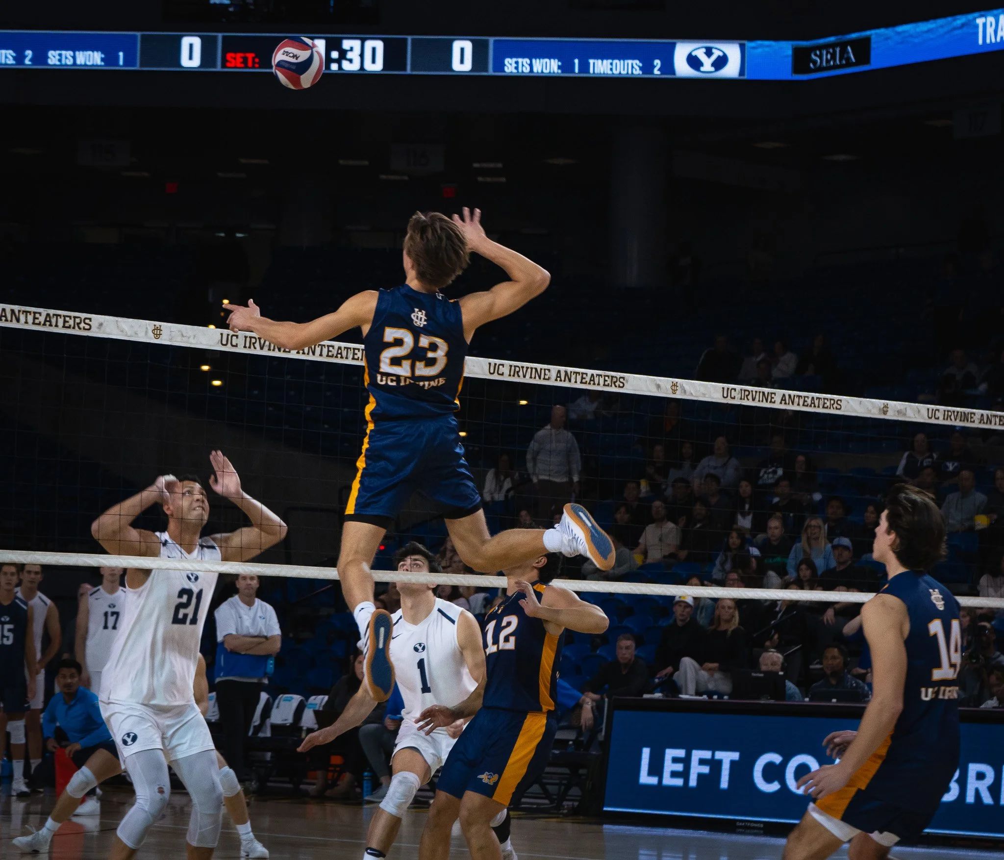 Volleyball match at UC Irvine Anteaters indoor gym, with players in blue and white uniforms jumping and preparing to block or spike the ball over the net, and spectators seated in the background.