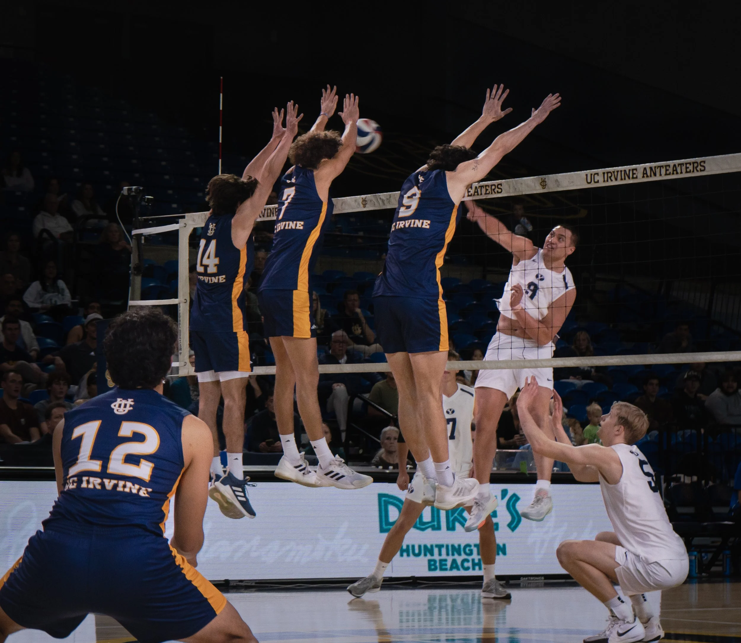 Volleyball players in action during a match, with three players from UC Irvine jumping to block the ball at the net, while a player from the opposing team prepares to spike the ball.