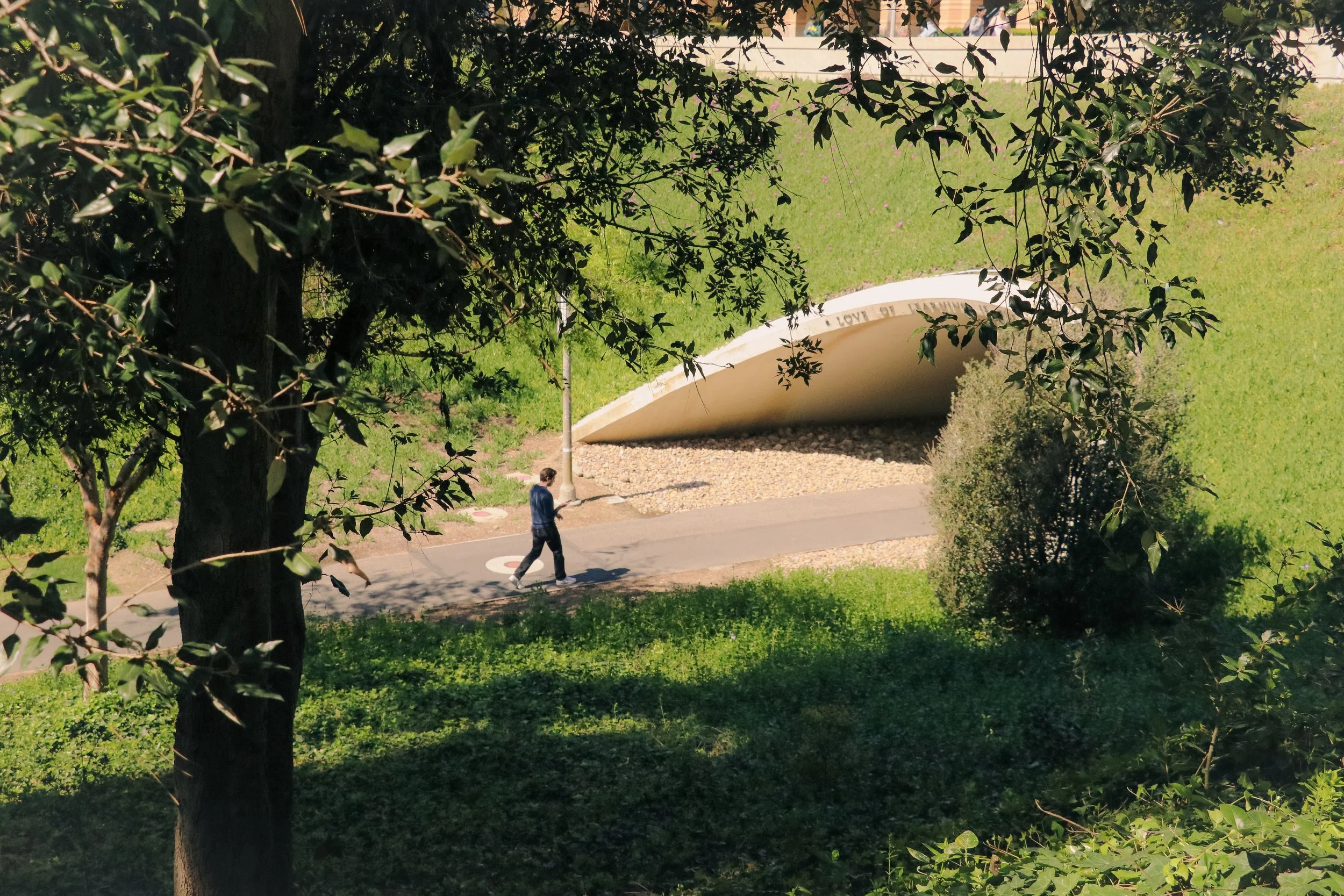 A person walking on a sidewalk past a unique, curved white structure with writings, in a park or green space, partly shaded by trees.