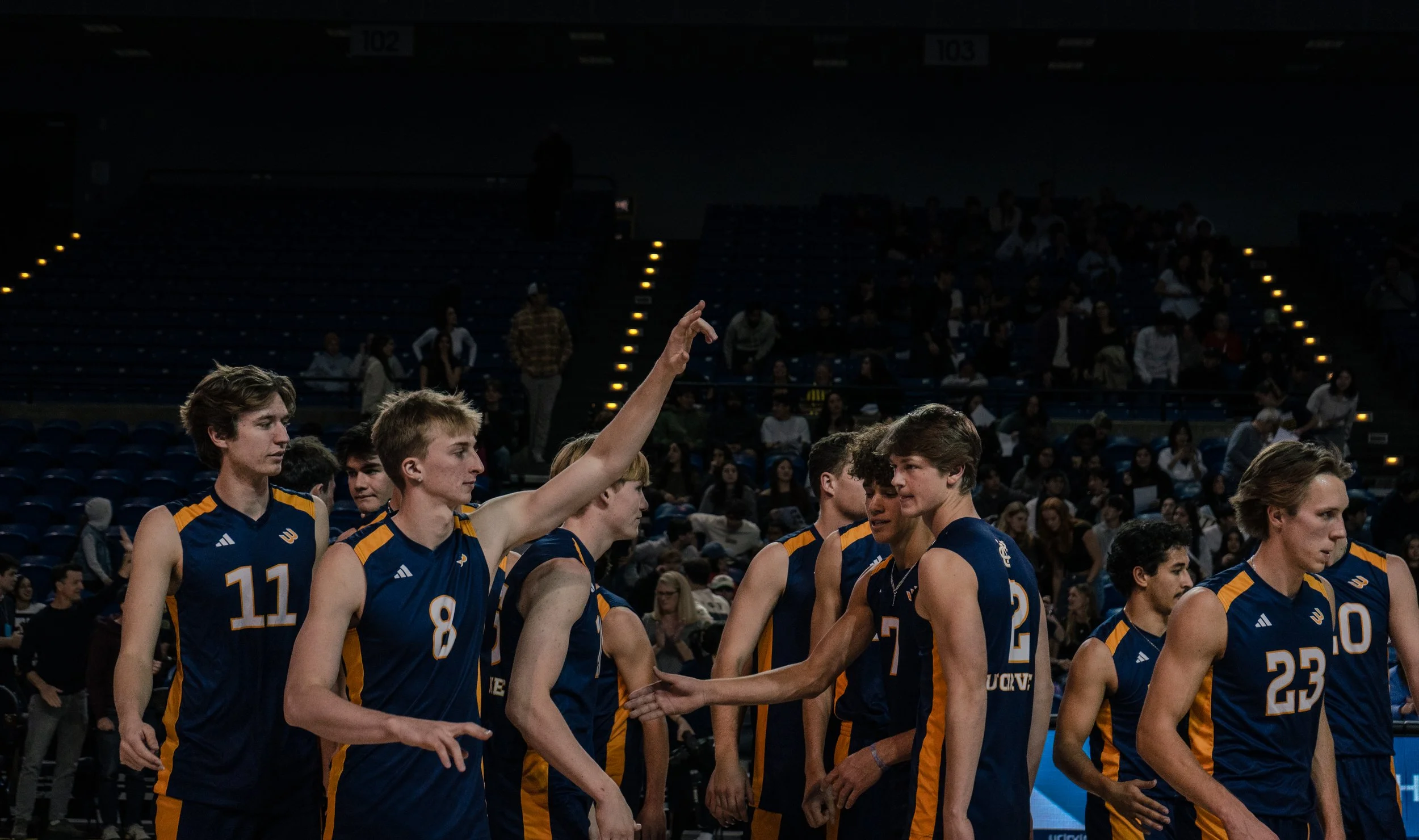 A group of male volleyball players in navy blue and orange uniforms on the court, with one player raising his arm in the air in a gymnasium filled with spectators.