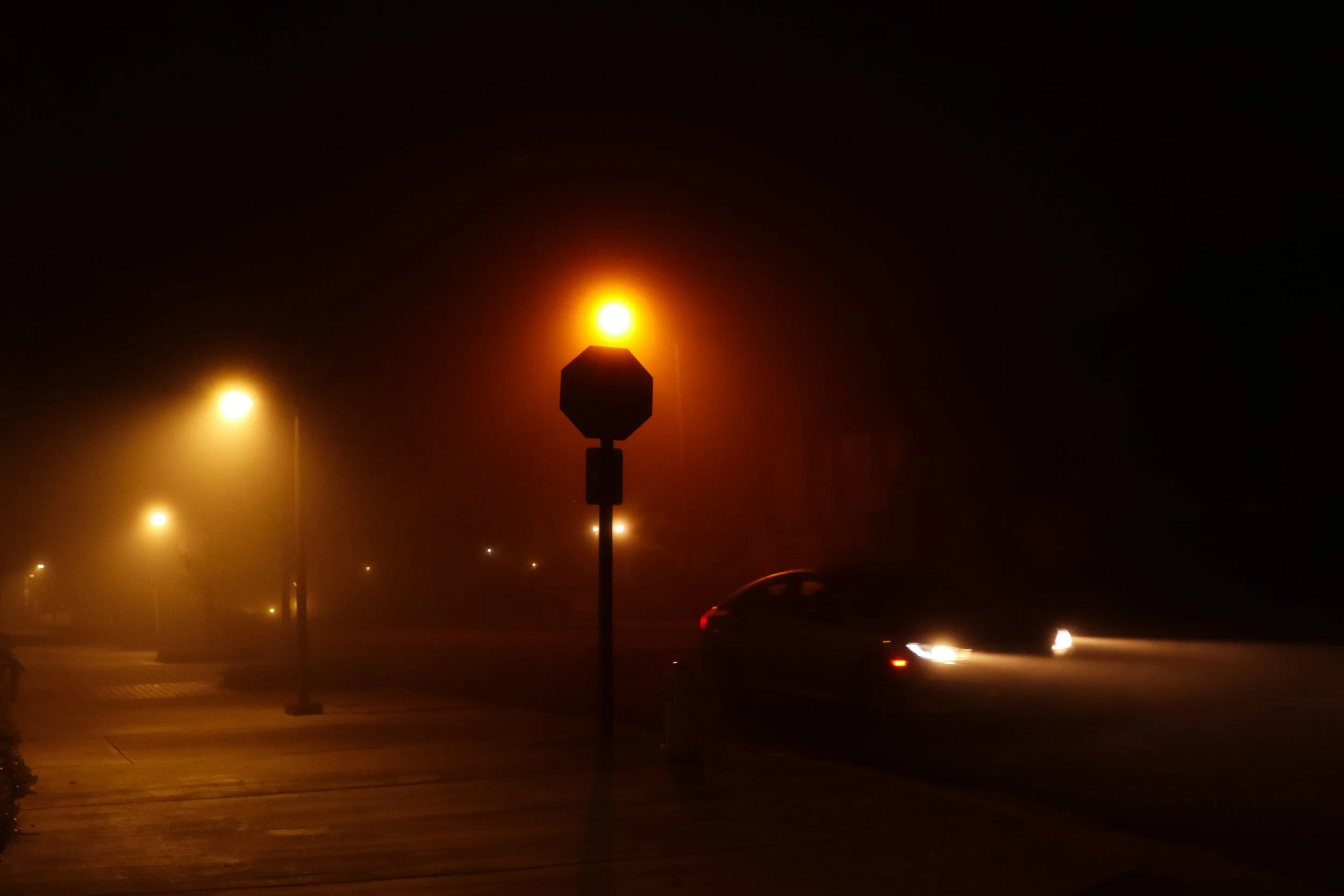 Nighttime scene with streetlights illuminating a foggy street, a stop sign, and a car with headlights on.