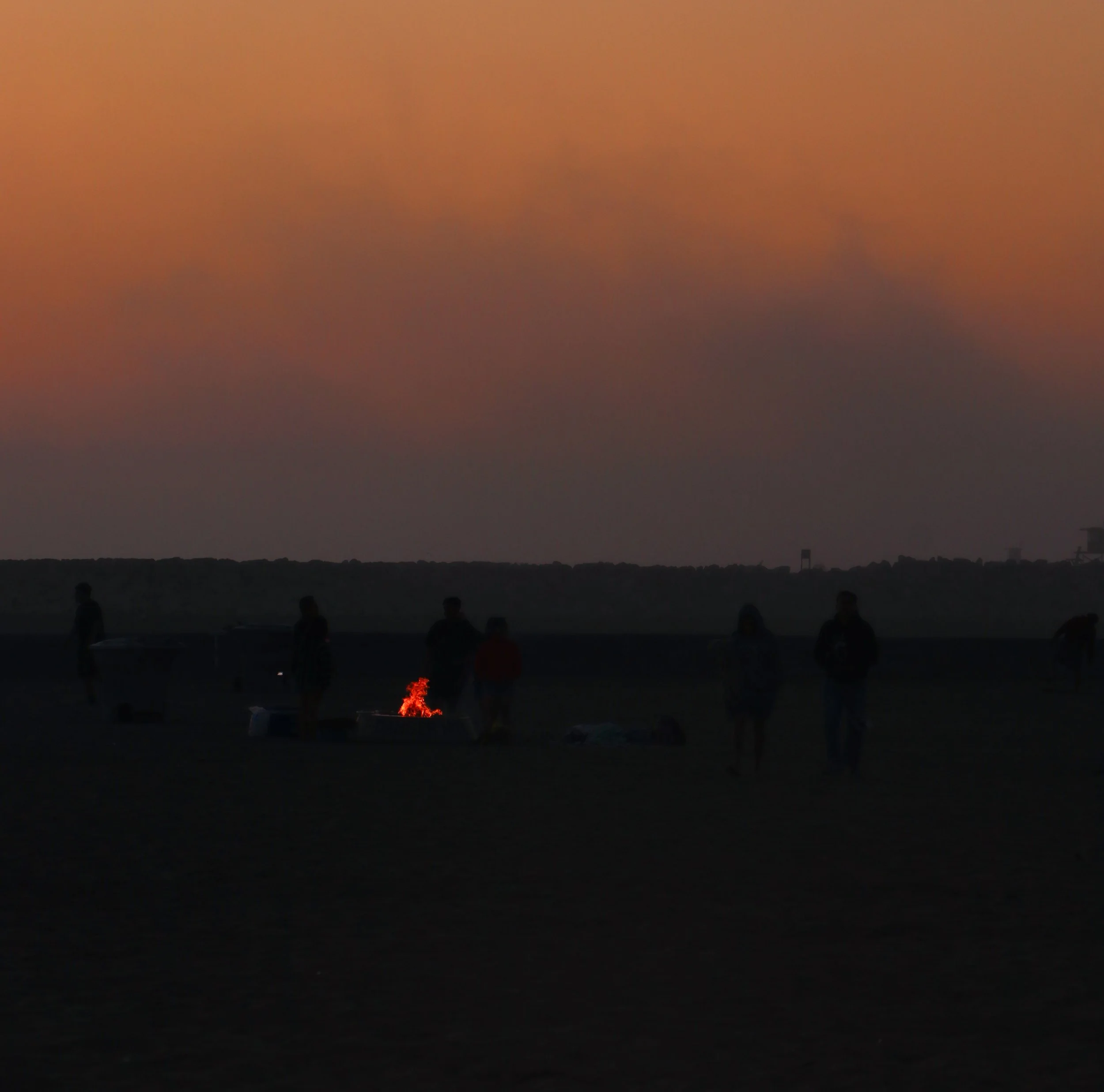Silhouettes of people standing on a dark beach at sunset, with a small fire burning and colorful clouds in the sky.