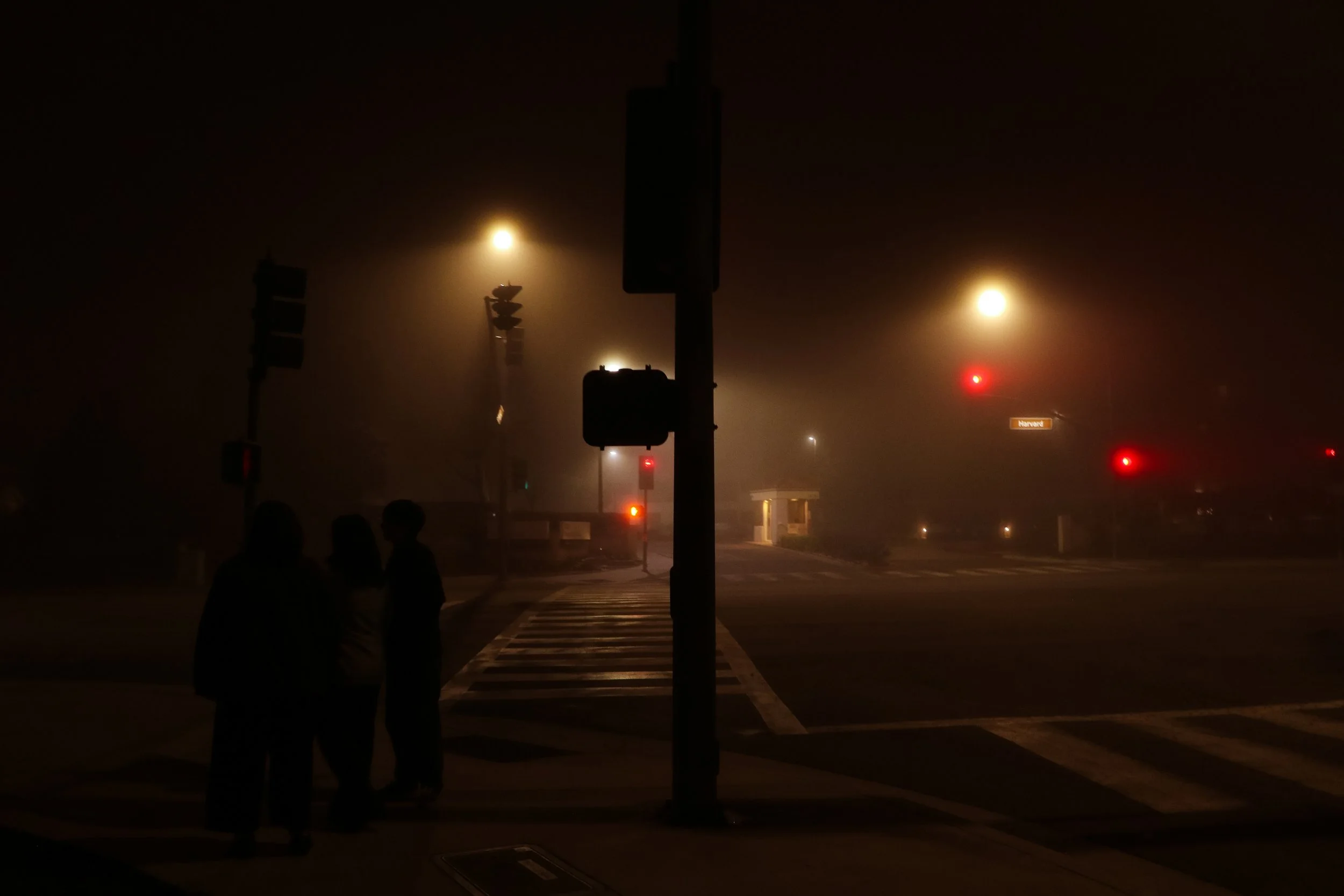 Three people in silhouette standing at a foggy, dimly lit street corner at night, illuminated by streetlights and traffic signals.