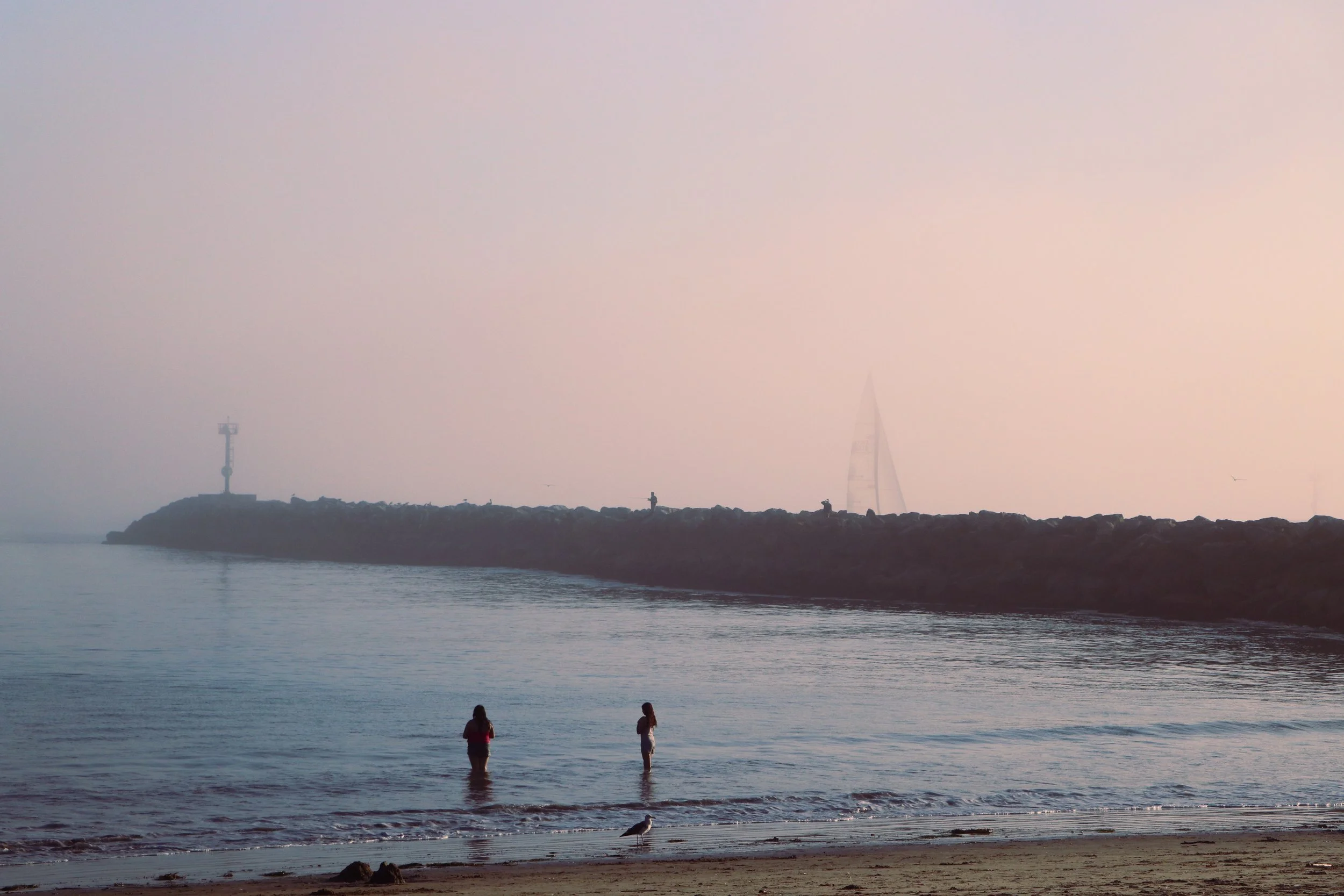 Two women standing in the ocean near a sandy beach during foggy weather, with a sailboat and a lighthouse on a breakwater in the background.