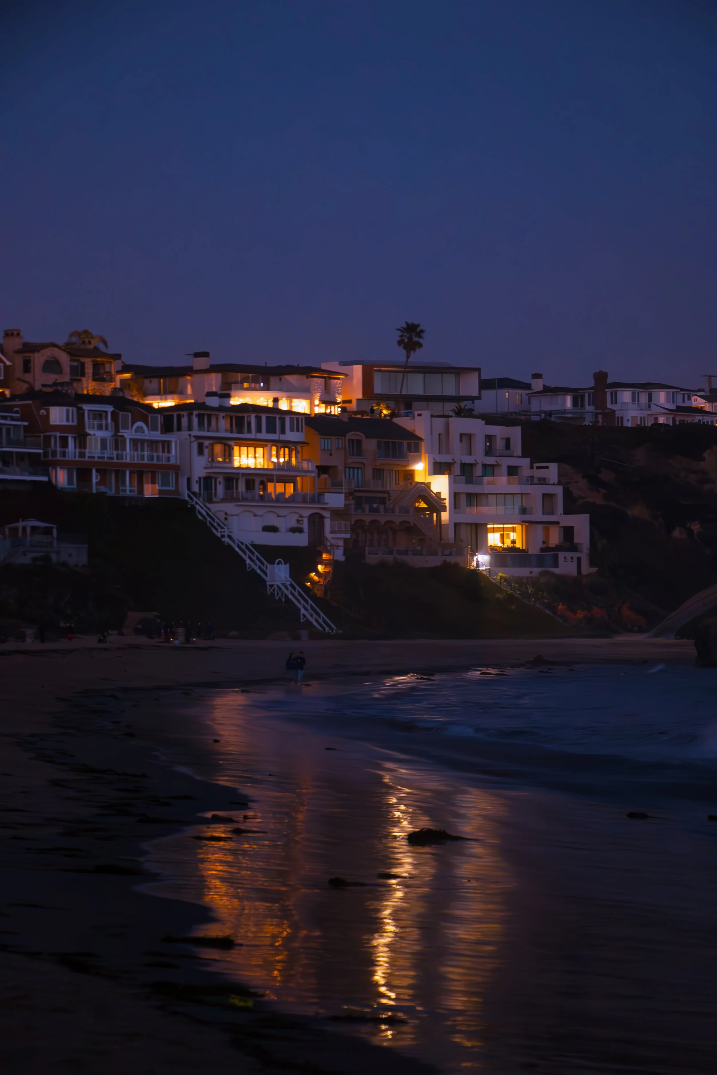 Beach at dusk with houses on cliffs illuminated by warm interior lighting, reflecting on the wet sand and gentle sea waves.