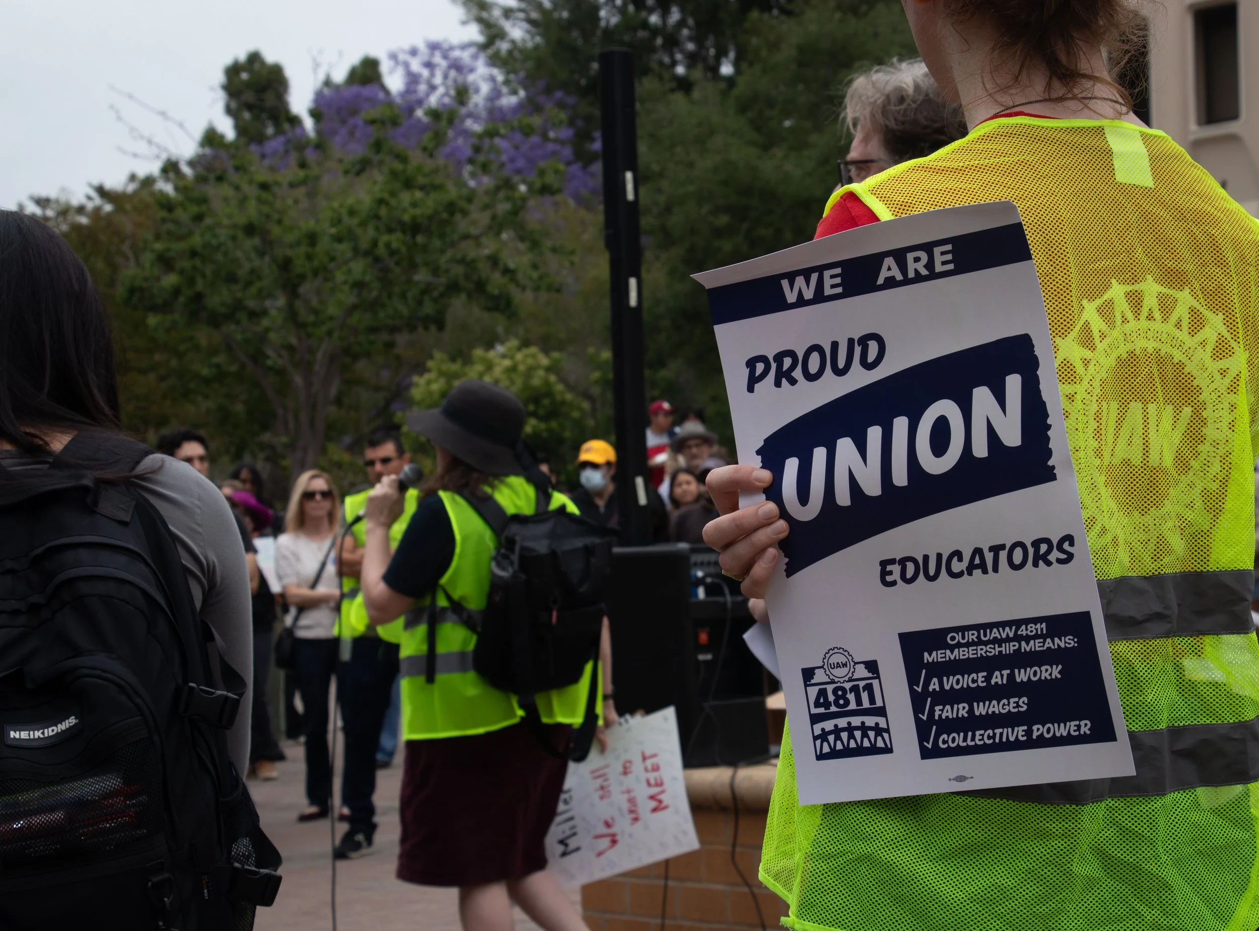 Protesters gather outdoors, wearing yellow vests. One protester holds a sign that reads 'We Are Proud Union Educators' with the UAW 4811 logo and message about union membership benefits.