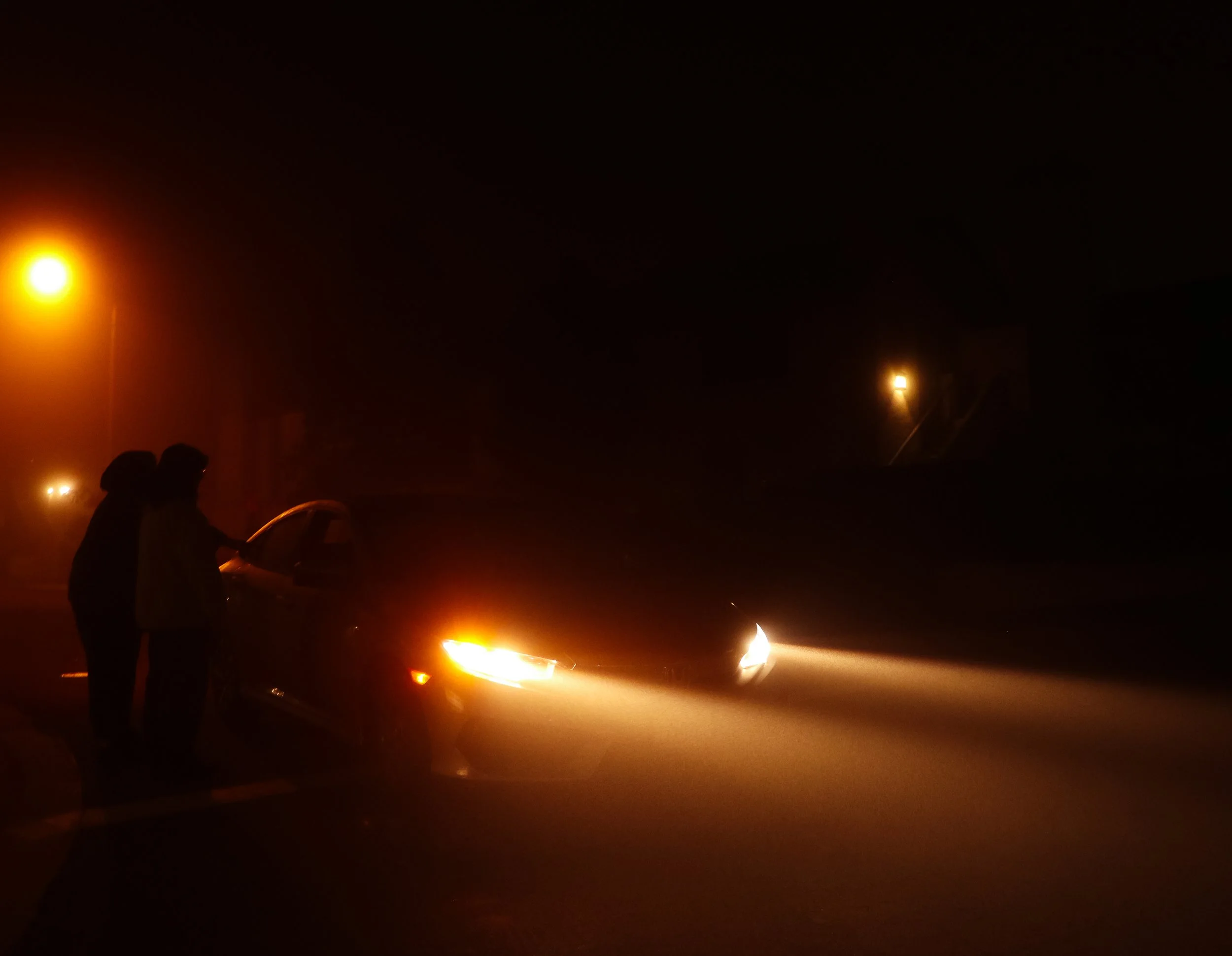 Two silhouetted individuals stand next to a car on a dark, foggy road illuminated by streetlights, with headlights on.