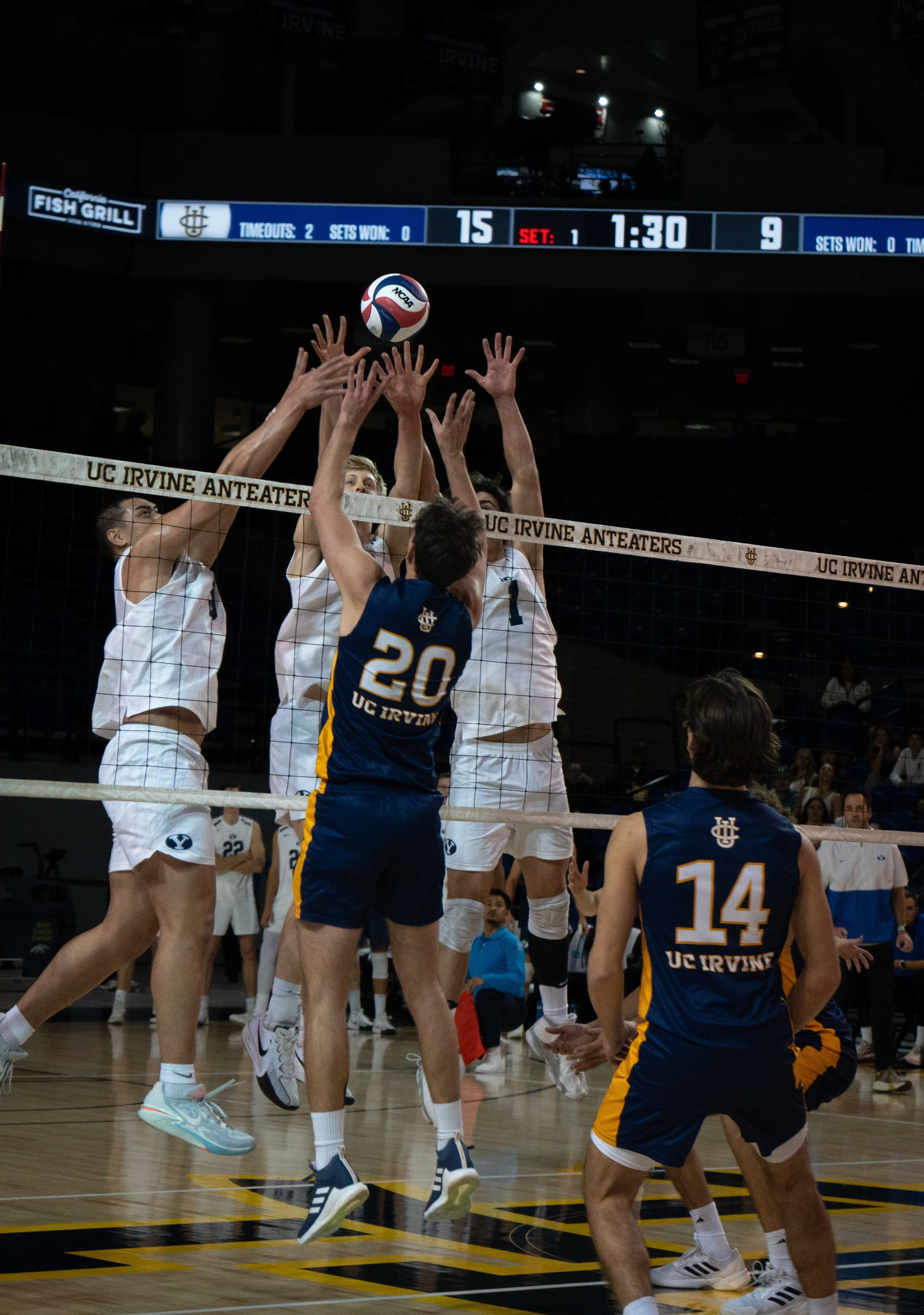 College volleyball game at UC Irvine, players jumping to block and spike the ball at the net.