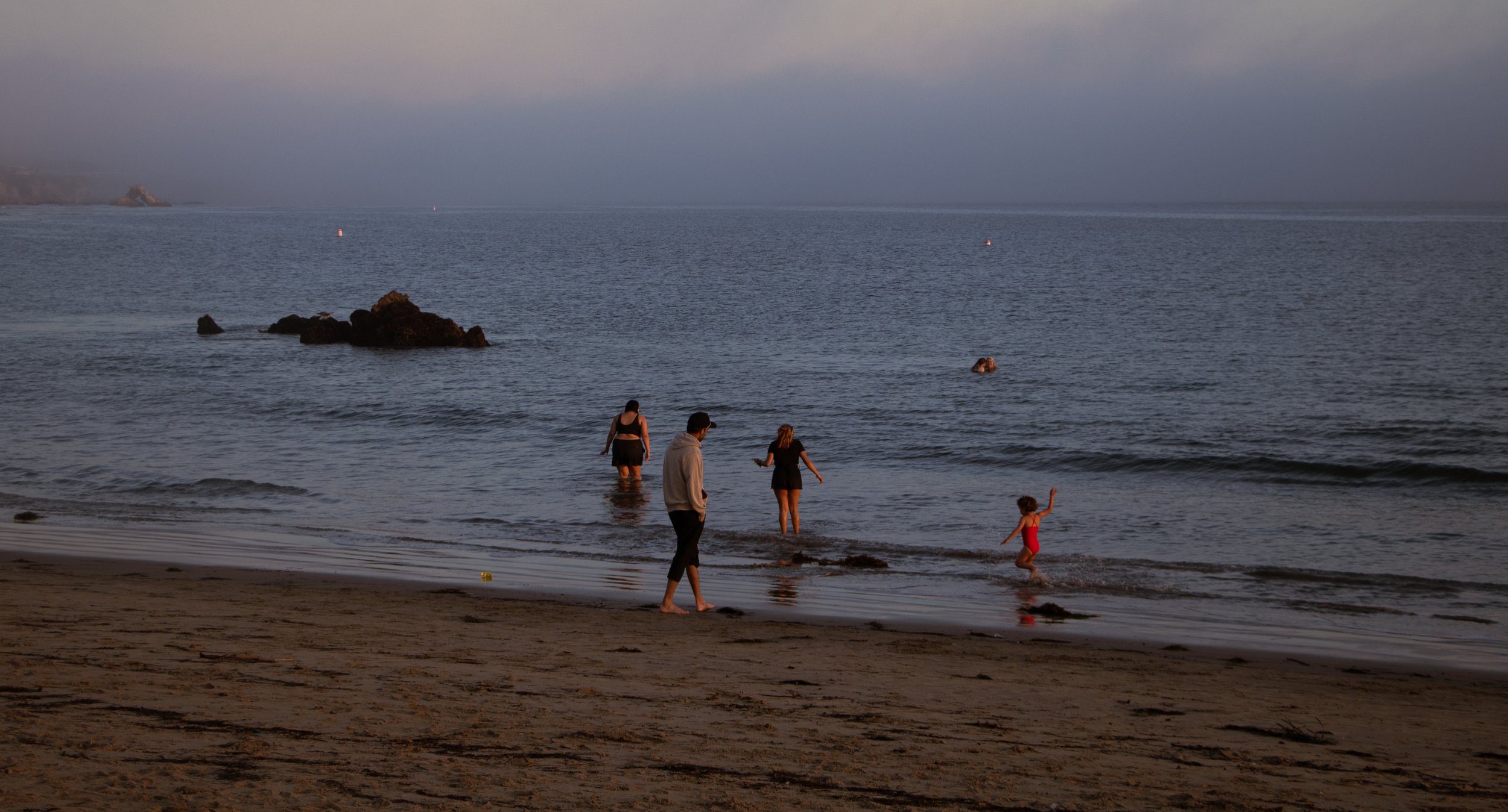 People at the beach, standing and playing in the shallow water, with rocks in the foreground and a calm ocean in the background during dusk.