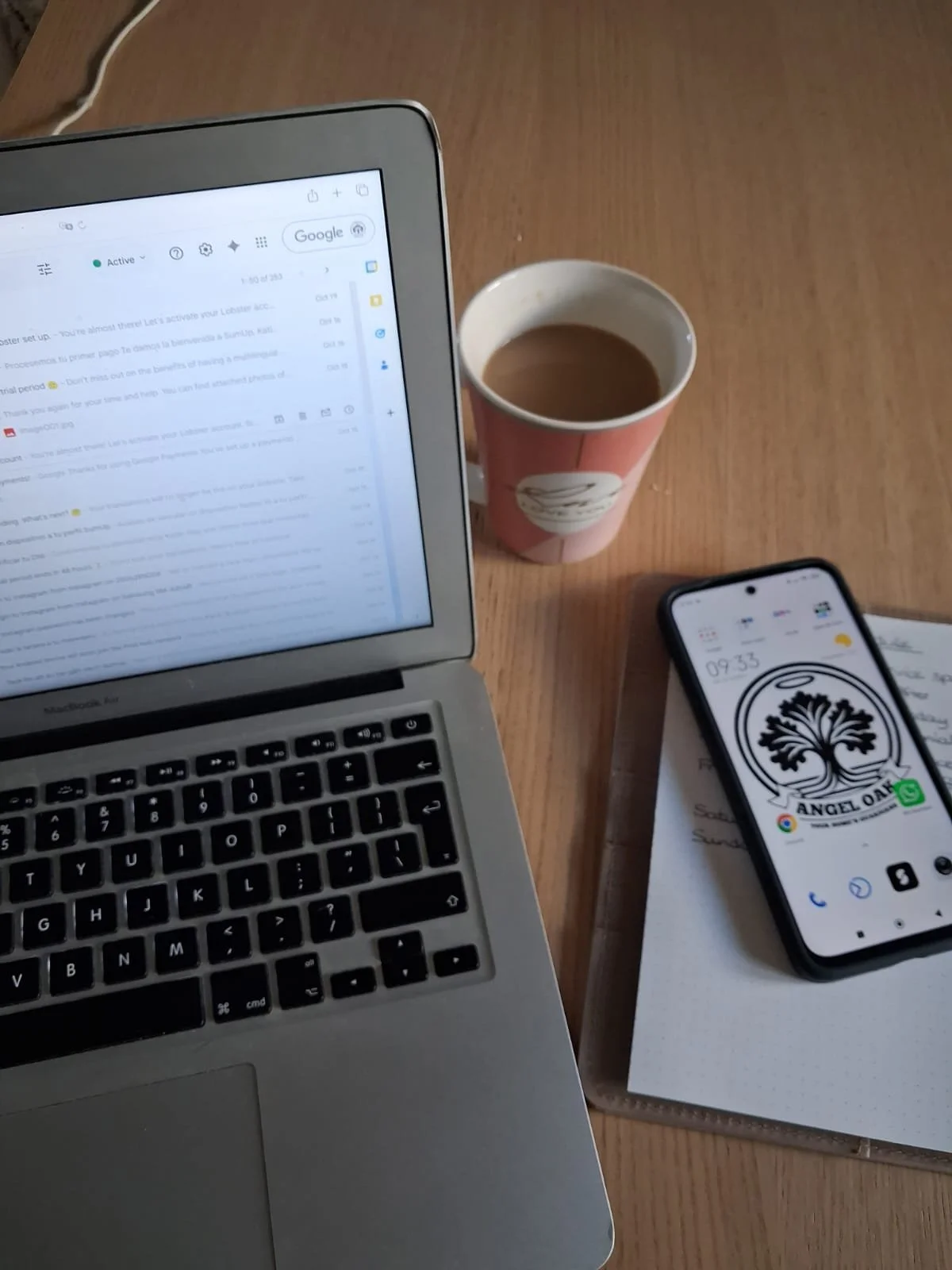 A laptop displaying an email inbox, a paper cup filled with coffee, a smartphone with a logo of a tree and the text "Angel Oak," and a notebook with handwritten notes on a wooden table.