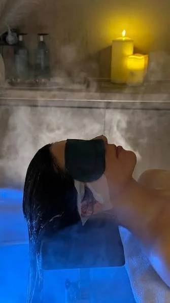 A woman receives a steam facial treatment in a spa, lying back with a towel over her face, bathed in blue and yellow lighting, with candles and toiletries in the background.