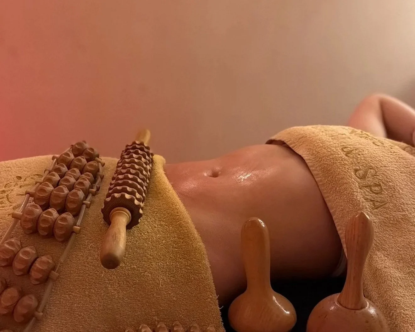 Person lying on a massage table receiving a hot stone massage, surrounded by various massage tools and hot stones, with a towel covering their torso.